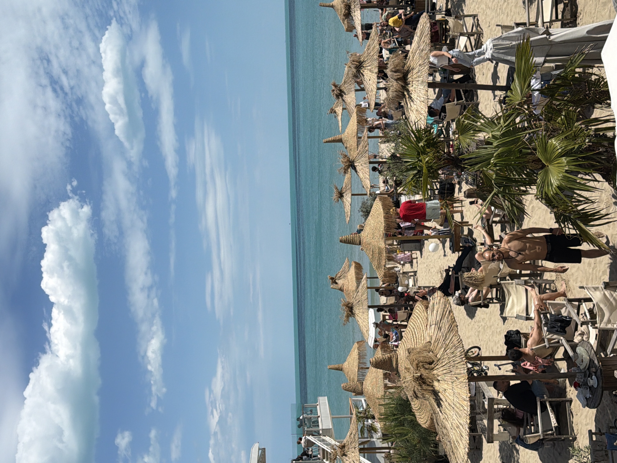 The image captures a vibrant beach scene in Varna, Bulgaria. The sky is predominantly blue with scattered cumulus clouds. The turquoise sea stretches to the horizon, separating the sky from the sandy beach. On the beach, numerous straw sun umbrellas are erected, providing shade for beachgoers. People are lounging on chairs and sunbeds, while others stroll along the shoreline. Palm trees and other beach flora are interspersed throughout the scene, adding a touch of tropical ambiance. A multi-level structure or building, possibly a bar or restaurant, is visible in the distance. The atmosphere suggests a relaxed and busy beach resort area.