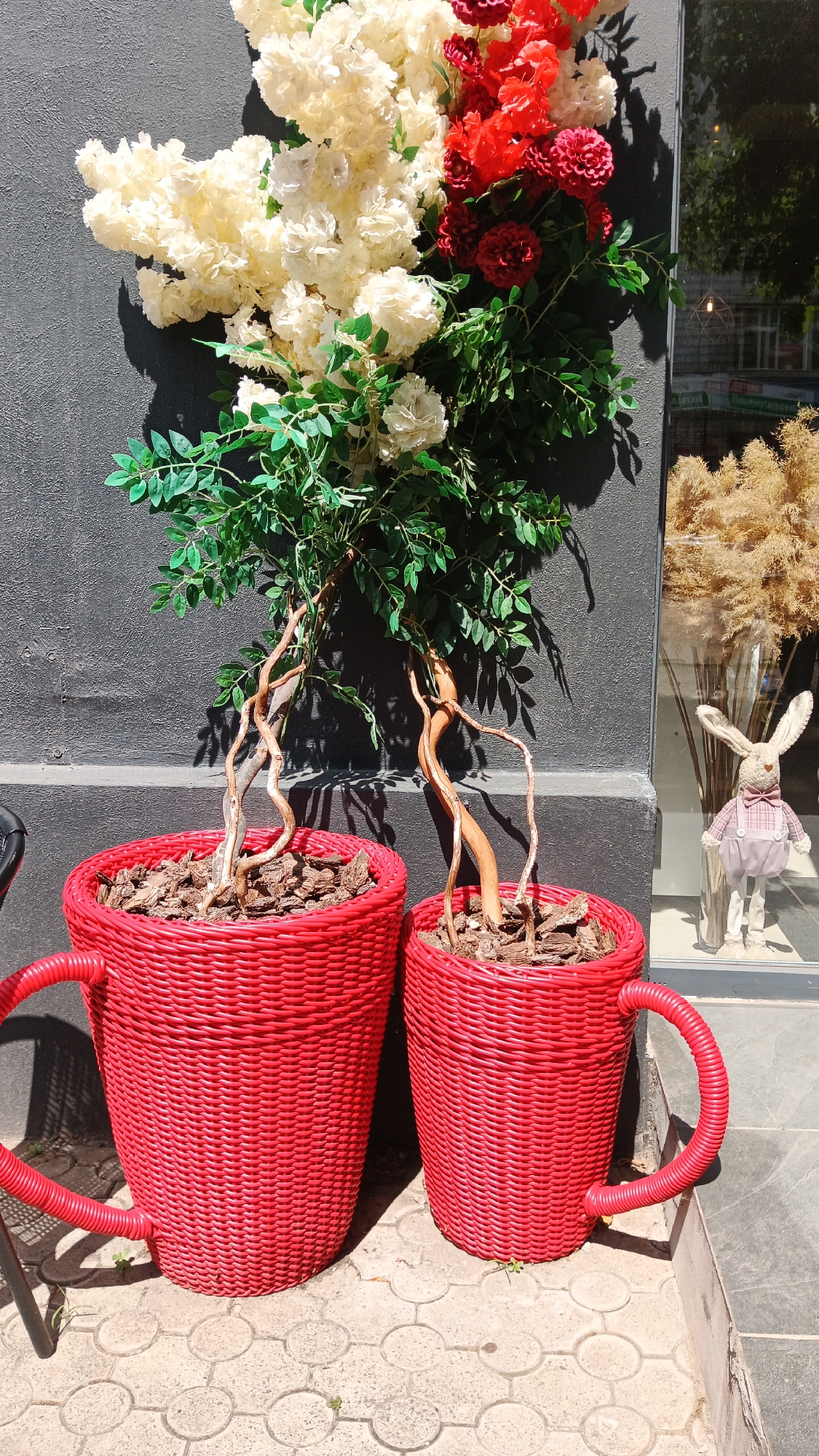 Two large, red, woven planters shaped like mugs are placed on a patterned sidewalk in front of a dark gray textured wall. The planters contain bonsai-like trees with intricate, twisted branches and green leaves. Above the planters, a decorative floral arrangement features white and red flowers mixed with greenery. To the right, a shop window is visible, reflecting the street and displaying a light-colored dried floral arrangement and a decorative bunny wearing a pink checkered outfit. The image appears to be taken in Kryvyi Rih, Ukraine, given the architectural context and typical urban environment.