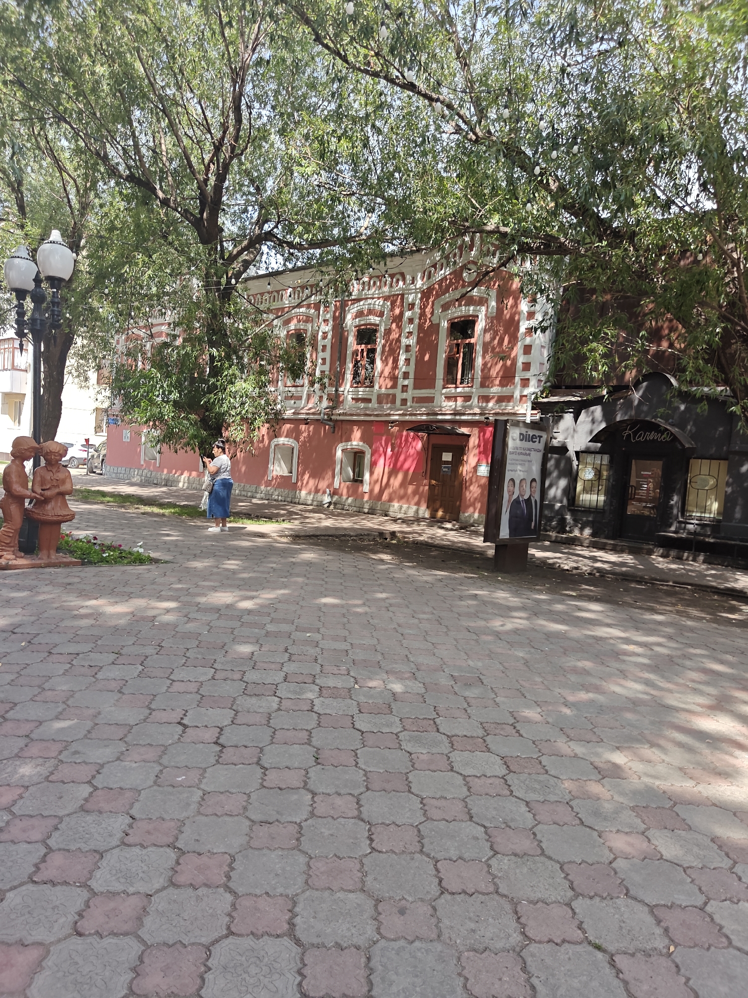 The image captures a sunny day in the city of Petropavl, Kazakhstan, on a street with ornate, pink-colored buildings. In the foreground, a patterned brick sidewalk is visible, casting dappled shadows from overhead trees. To the left, two child-like statues stand near a lamppost. A woman in a striped shirt and a blue skirt is standing on the sidewalk, holding a smartphone and appearing to take a photo. Behind her, a vibrant pink building with decorative white trim and arched windows dominates the mid-ground. Further to the right, a darker building with a sign that reads "Karma" is partially obscured by trees. An advertising billboard stands on the sidewalk between the pink building and the "Karma" establishment. The scene depicts a typical urban streetscape with architectural elements and pedestrian activity.