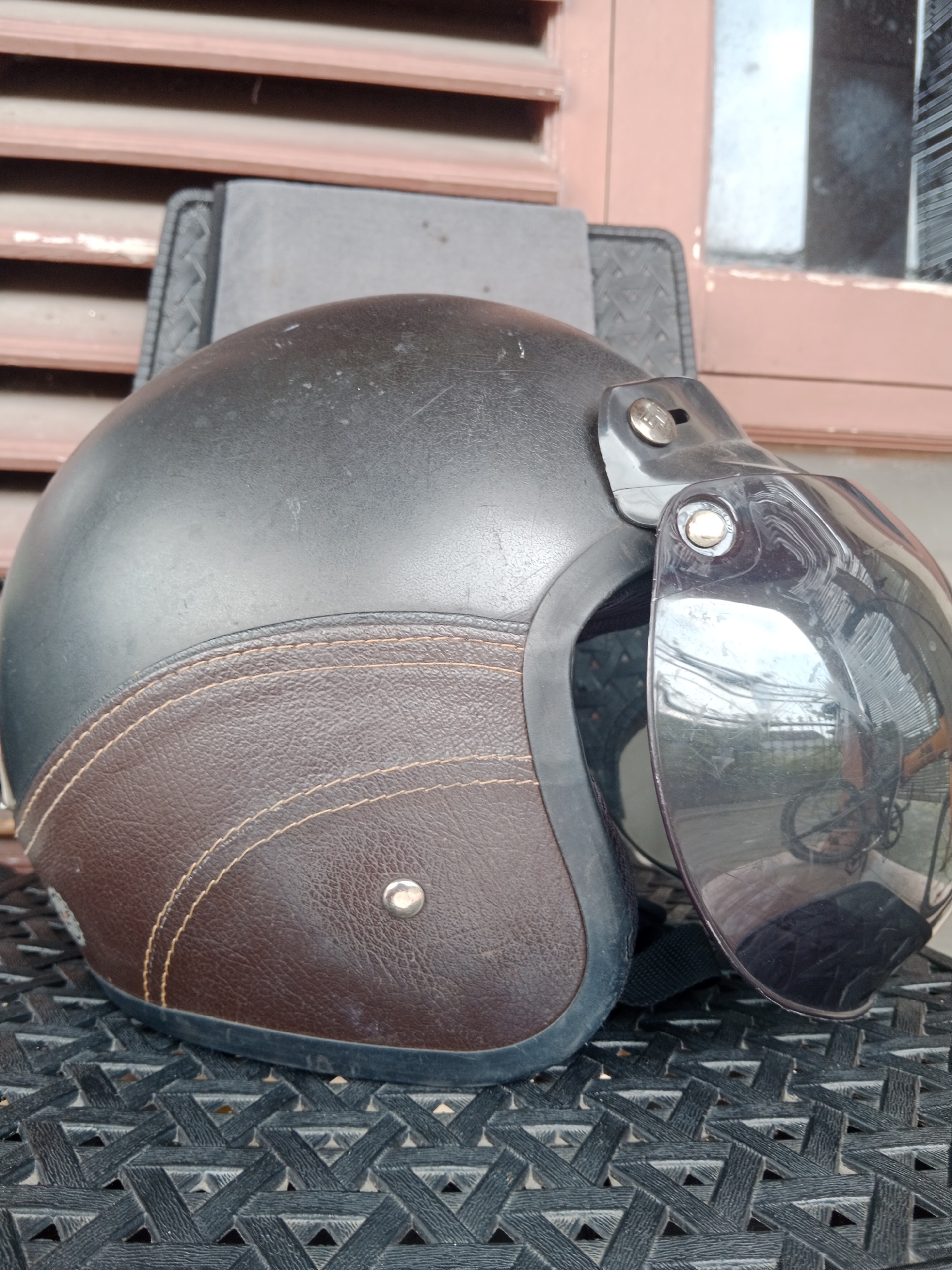 A black motorcycle helmet with a brown leather accent is displayed on a black woven surface, likely a chair or mat. The helmet features a clear bubble visor, reflecting the surroundings which appear to be outdoors with sunlight. Behind the helmet, a slatted wooden structure and a window with reflections are visible. The scene is likely in Banguntapan, Indonesia, based on the visual cues of the architecture and common outdoor elements.