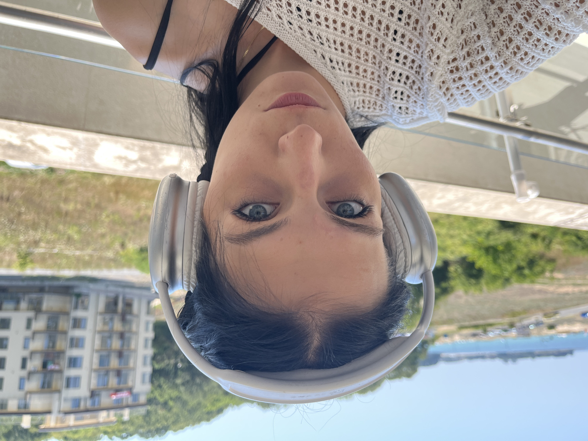 A close-up, eye-level, selfie-style, medium shot captures a young woman with long, dark hair wearing silver over-ear headphones. She is looking directly at the camera with a neutral expression. Her face is illuminated by natural light, and she appears to be outdoors. She is wearing a white, crocheted off-the-shoulder top. In the background, a light-colored, multi-story building with balconies is visible, set against a backdrop of green trees and a bright blue sky. To the left, a body of water can be seen with a beach and a road. The scene appears to be in Kiten, Bulgaria, likely on a balcony overlooking the coastline.