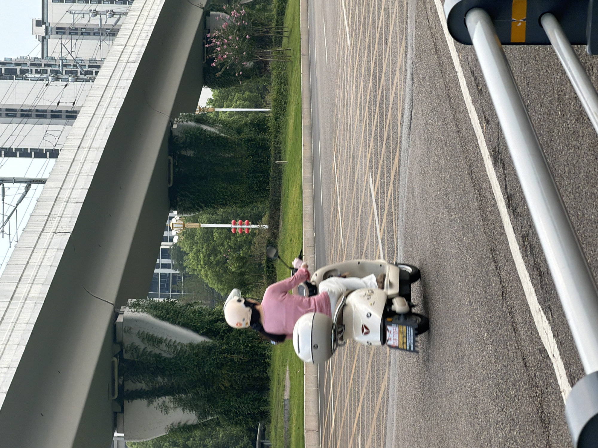 A woman wearing a pink long-sleeved shirt, white pants, and a light-colored helmet rides a light-colored scooter on an asphalt road. The scooter has a white storage box on the back and a license plate with Chinese characters and numbers. The road has yellow grid markings and a white lane line. In the background, a concrete overpass with visible cracks runs across the frame, supported by pillars covered in greenery. Beyond the overpass, modern buildings rise into the sky, and utility wires are strung between poles. Trees and bushes line the side of the road. The image appears to be taken outdoors during the daytime. The license plate on the scooter is identified as belonging to Jiangjin, China.