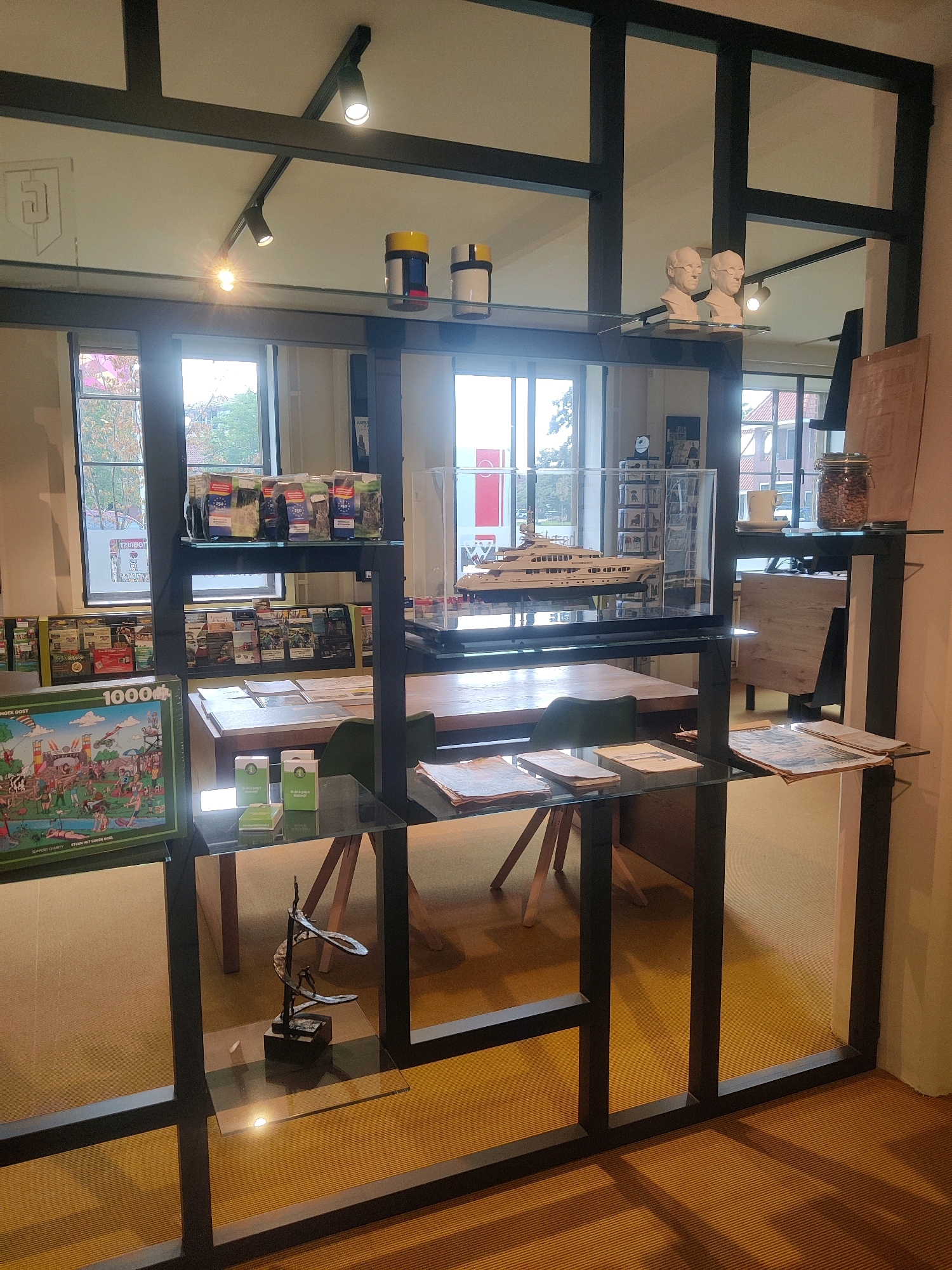 The image depicts the interior of a retail establishment, possibly a gift shop or museum store, located in Winterswijk, Netherlands. The space is organized with shelving units and display cases. On the upper shelves, two busts of men wearing glasses are displayed, alongside cylindrical objects with yellow and blue detailing. Below, a model of a white yacht sits inside a glass case. Further down, there are shelves stocked with various items, including puzzles, packaged goods, and magazines. A glass table with some papers and small boxes is visible on a lower shelf. In the foreground, a metal sculpture is displayed on a glass shelf. The environment features warm lighting and a textured floor.