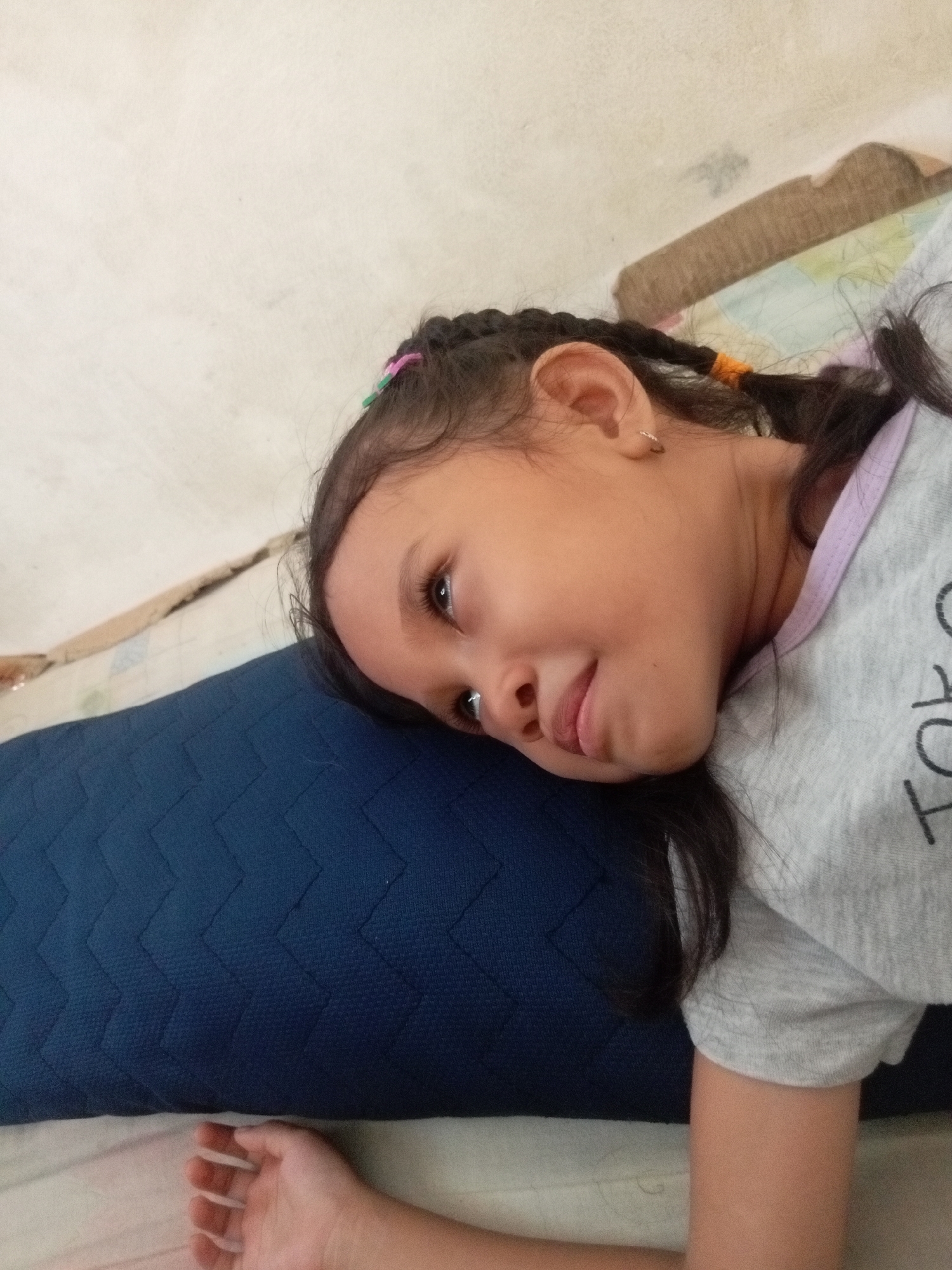 A young girl with braided dark hair, adorned with pink and green elastics, lies on her side resting her head on a dark blue quilted pillow. She wears a grey t-shirt with purple trim and a partial graphic print. Her eyes are open, looking towards the viewer with a slight smile. Her left arm is visible beneath the pillow, with fingers slightly curled. The background consists of a textured off-white wall and a light-colored mattress or bedspread. The scene appears to be an indoor setting, likely a bedroom. The image does not provide any specific details to determine the exact location within Batam, Indonesia.