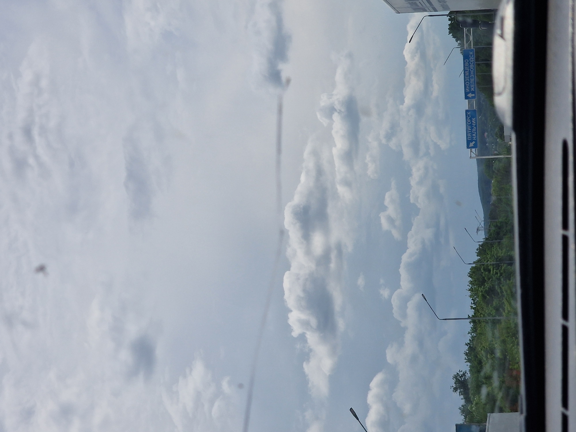 This is a photograph taken from inside a vehicle, looking out at a cloudy sky and a landscape with trees and signs. The signs indicate directions to "ПЯТИГОРСК НАЛЬЧИК" (Pyatigorsk, Nalchik) and "ИНОЗЕМЦЕВО ЖЕЛЕЗНОВОДСК" (Inozemtsevo, Zheleznodorozhny). This indicates the location is near the crossroads for these cities, consistent with the area around Змейка, Russia, which is a rural locality within the city of Zheleznodorozhny. The scene is a roadside view, with streetlights visible along the road and a dense green tree line in the middle ground. The sky is predominantly filled with white and grey clouds. There are no people or overt actions depicted in the image.