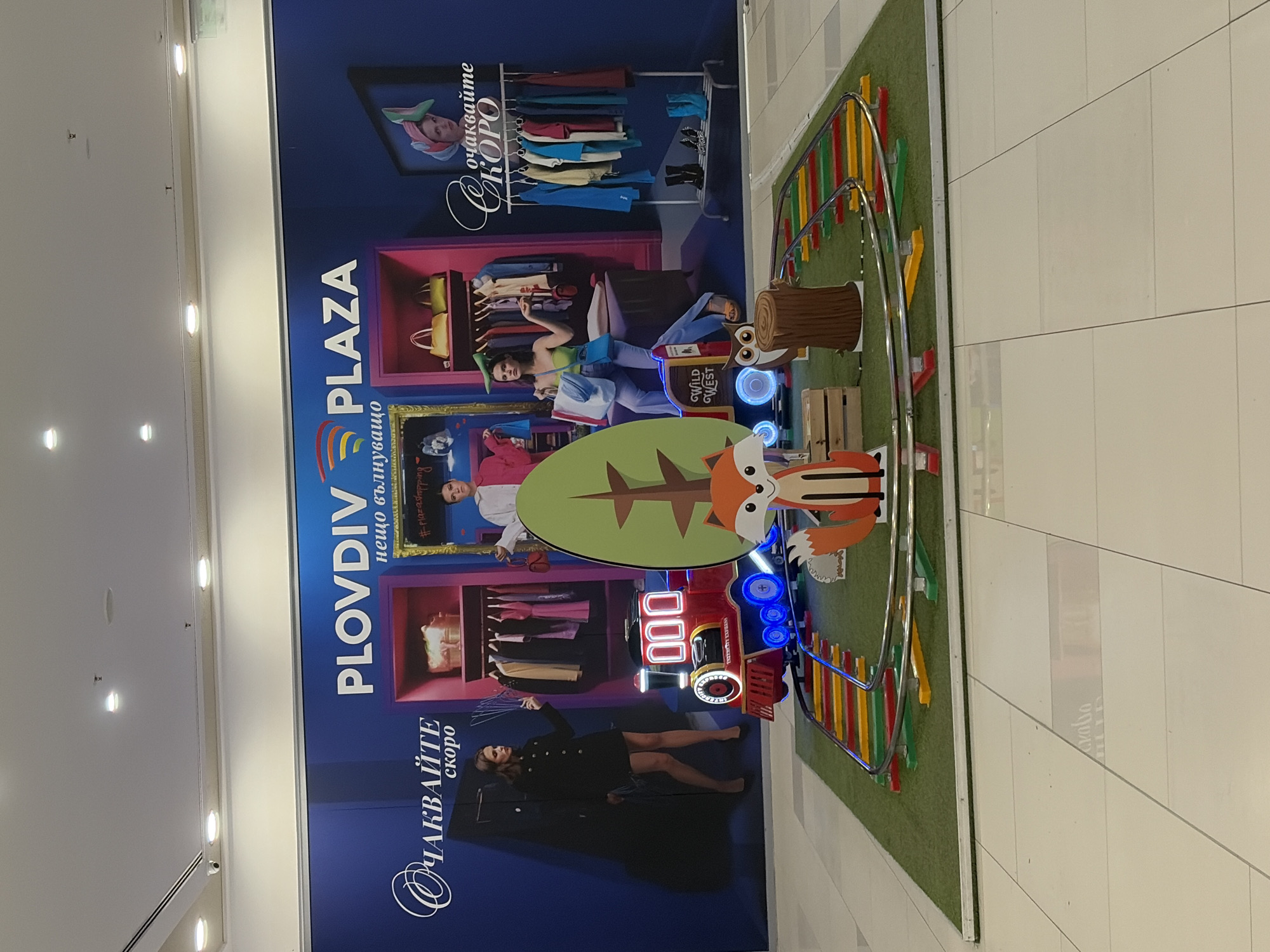 The image shows an advertisement for a shopping center in Plovdiv, Bulgaria named "PLOVDIV PLAZA". The advertisement is on a wall and features images of people and clothing, as well as the text "ОЧАКВАЙТЕ скоро" and "нещо вълнуващо". There is also a display on the floor in front of the advertisement, which includes a wooden fox and owl, a toy train set, and a green patch of artificial turf.