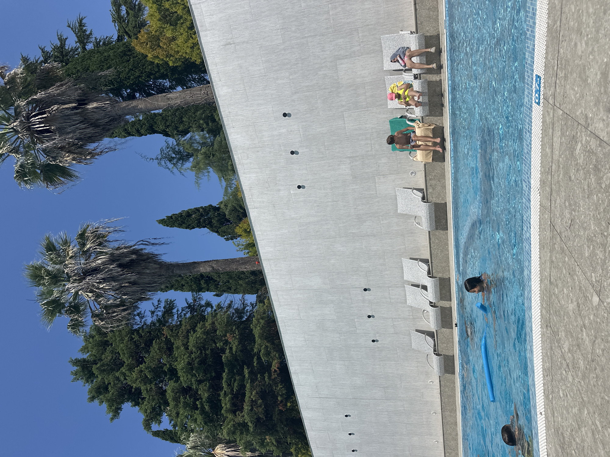 An outdoor swimming pool is depicted under a clear blue sky, surrounded by lush green trees and tall palm trees, suggesting a warm climate. The pool is filled with bright blue water, and a few people are visible enjoying their time. In the foreground, a young boy is swimming, and a young girl is in the pool with a blue foam noodle. In the background, there are several lounge chairs lined up against a grey tiled wall. Three children are seated on these chairs. One boy is wearing striped swim shorts, another child is wearing a yellow swimsuit and a pink hat, and a third child is wearing a grey hooded towel. The wall has black circular fixtures. The location is identified as Sochi, Russia, a popular resort city.