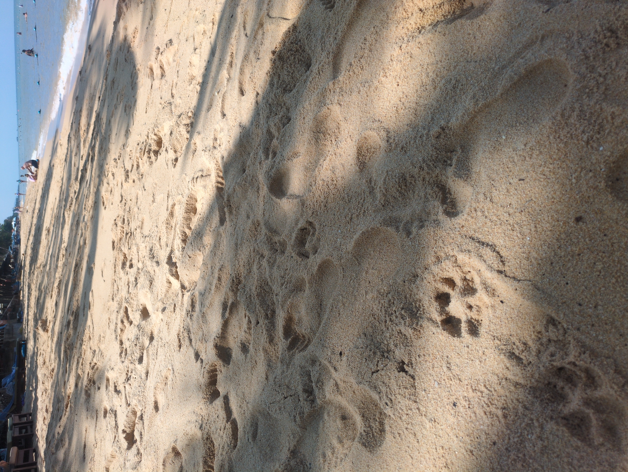 A beach scene at Phú Quốc, Vietnam. The sand is light beige, covered in footprints of varying sizes. A shadow stretches across the sand. The sea is visible in the background, with a few people in the distance.