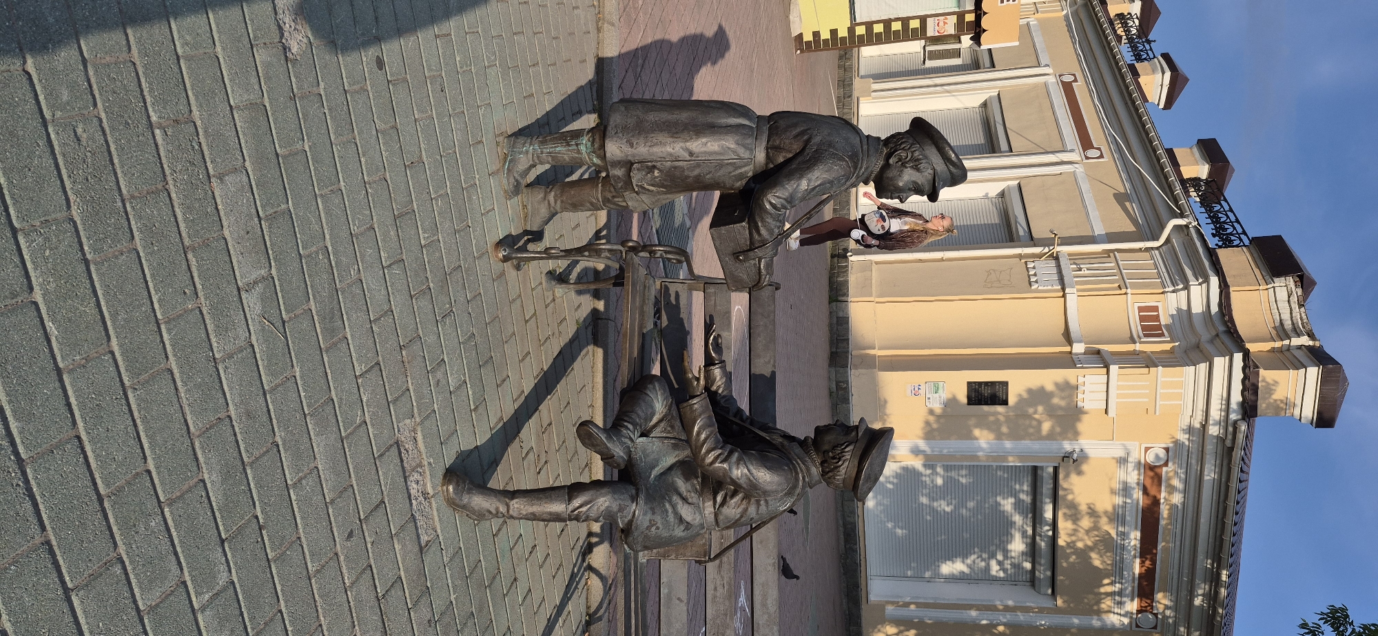 In Ussurijsk, Russia, a bronze sculpture depicting two young men in uniform is positioned on a paved sidewalk. The sculpture features one individual standing and holding a box, while the other is seated on a bench, facing the standing figure. A young woman, dressed in casual clothing, walks in the background behind the sculptures. The scene is set against a backdrop of a yellow building with architectural embellishments and closed shutters. The ground is covered in grey paving stones, and shadows are cast by the sculptures and a nearby tree. The sky is clear and blue.