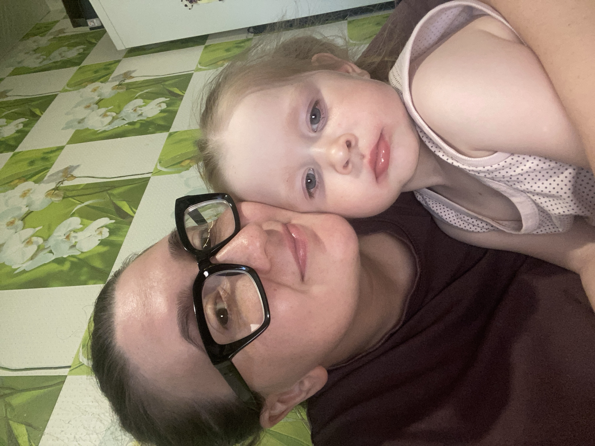 A woman with dark hair pulled back and large black-framed glasses smiles slightly, her face close to the camera. She is holding a young child, who looks directly at the camera with a neutral expression. The child has fair hair and blue eyes. They are both wearing casual clothing. The background is a wall with a green and white checkered wallpaper featuring white floral designs. The scene appears to be indoors, likely in a residential setting. The overall impression is of a candid selfie taken in a home environment.