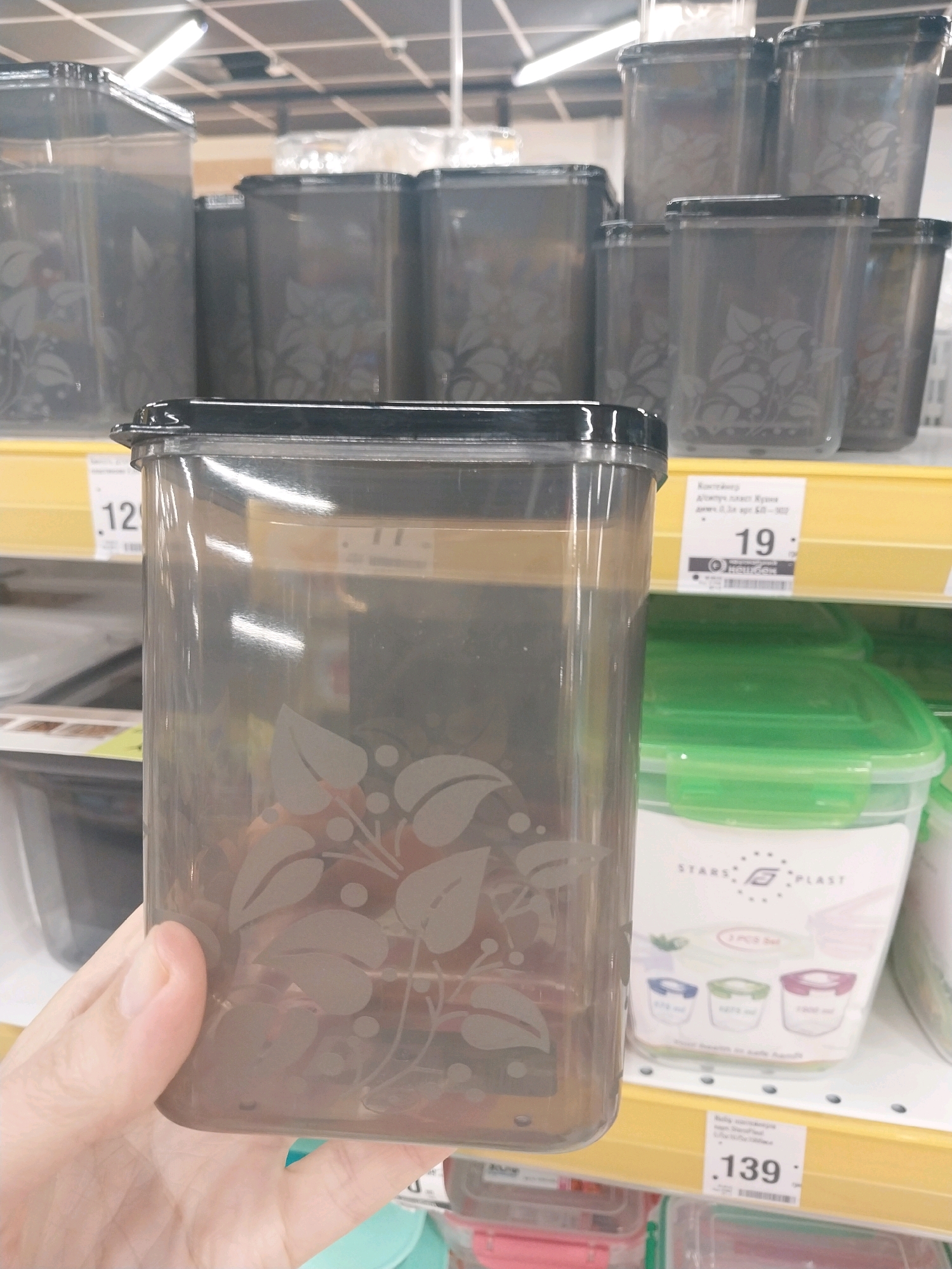 The image shows a close-up of a person's hand holding a transparent, rectangular plastic container with a leaf pattern. The container has a black lid. In the background, there are shelves displaying various plastic storage containers, some with green lids. Price tags are visible on the shelves, with one price tag showing "19" and another showing "139". The environment appears to be a retail store, likely a supermarket or household goods store. The lighting suggests an indoor setting. The image does not contain any specific information about the city of Dnipro, Ukraine.