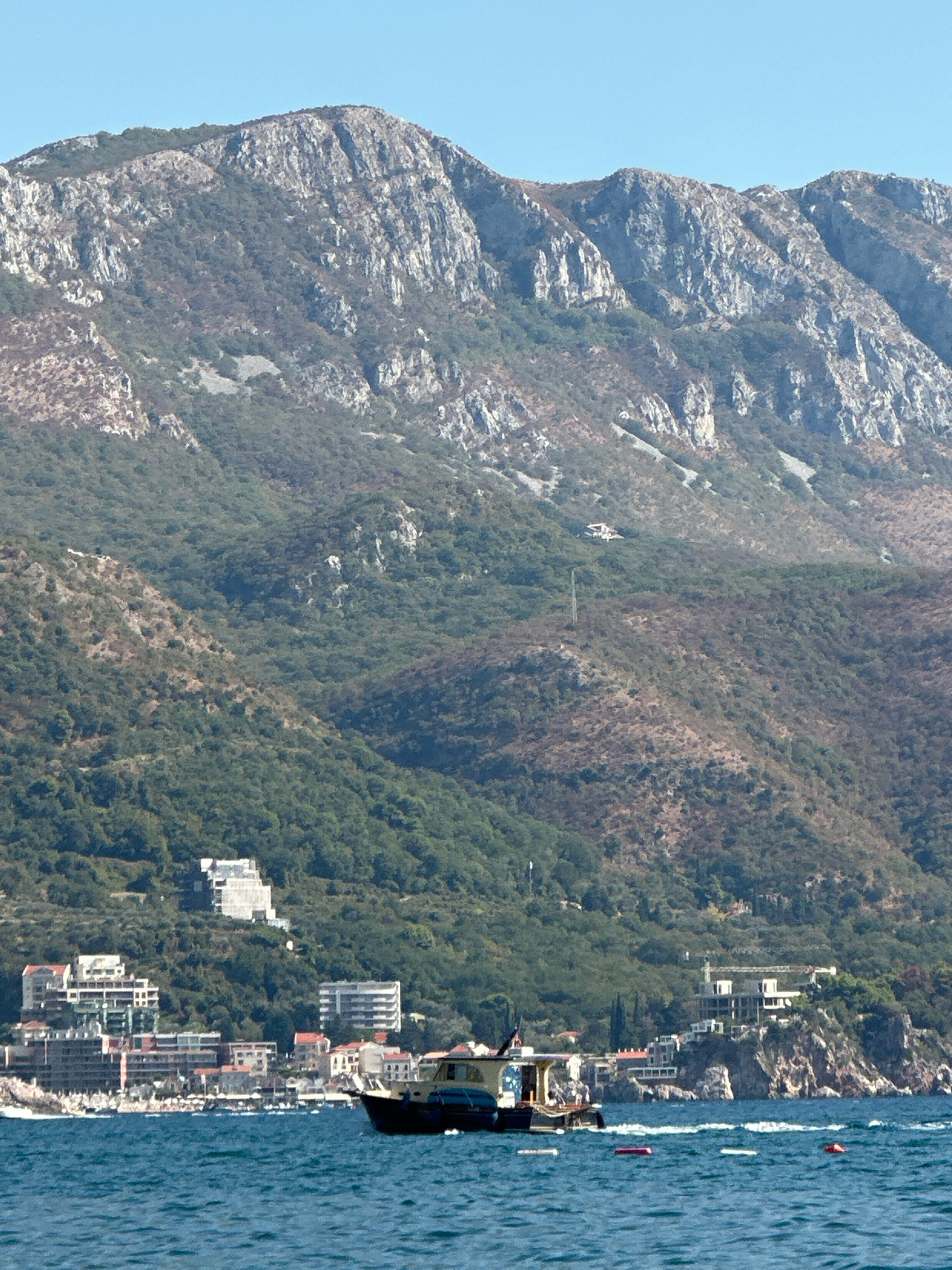 An aerial view captures the coastline of Boreti, Montenegro, under a clear blue sky. In the foreground, the cerulean waters of the Adriatic Sea are dotted with a few buoys. A dark-hulled boat with a light-colored cabin sails on the water, its stern facing the viewer.
The middle ground showcases the coastal town of Boreti, characterized by a cluster of buildings with white, beige, and red facades, some of which are modern multi-story structures and others appear to be older residential buildings. The town is nestled against a steep, densely forested mountainside. A few buildings are visible further up the slopes, suggesting development extending into the higher elevations. One building, a large white structure with a distinctive tiered design, is prominently situated on the hillside.
The background is dominated by a rugged, imposing mountain range. The upper slopes are rocky and barren, with stark white cliffs contrasting against patches of scrub and scattered green vegetation. The lower slopes of the mountains are covered in a thick, dark green forest, with visible valleys and ravines. The terrain appears steep and challenging, indicative of a Mediterranean mountain landscape. The overall scene depicts a scenic coastal area where human habitation meets a dramatic natural environment.