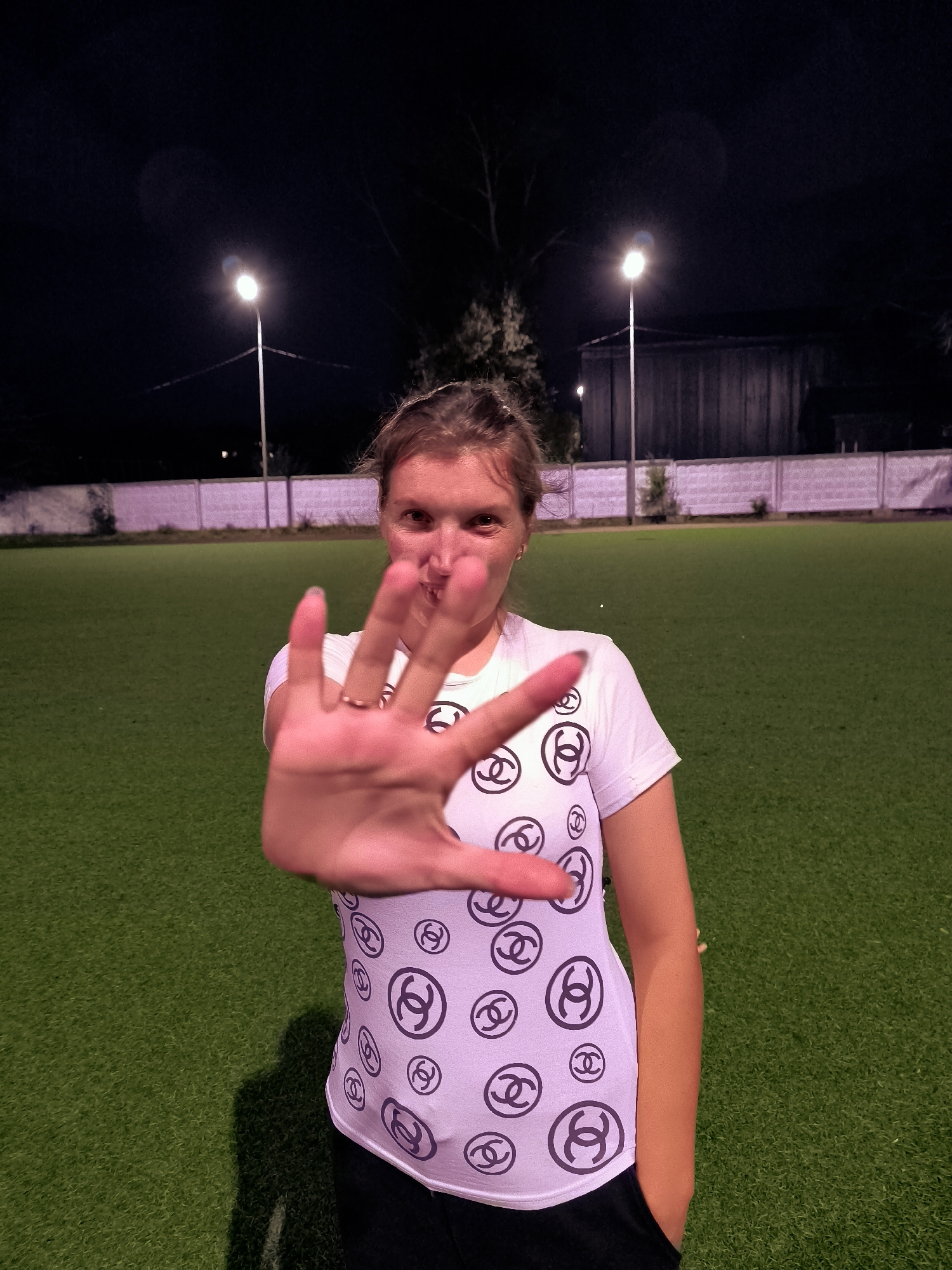 A woman stands on a bright green artificial turf field at night. She is wearing a white t-shirt with a repeating pattern of black Chanel logos. Her right hand is extended towards the camera, obscuring a portion of her face. She is looking at the camera with a slight smile. Behind her, two streetlights illuminate the field, casting long shadows. A white fence runs along the background, separating the field from a building. The overall mood is casual and playful, perhaps a candid shot taken during an evening outing. The time is night, and the weather appears clear. The location is Qarağandı city administration, Kazakhstan.