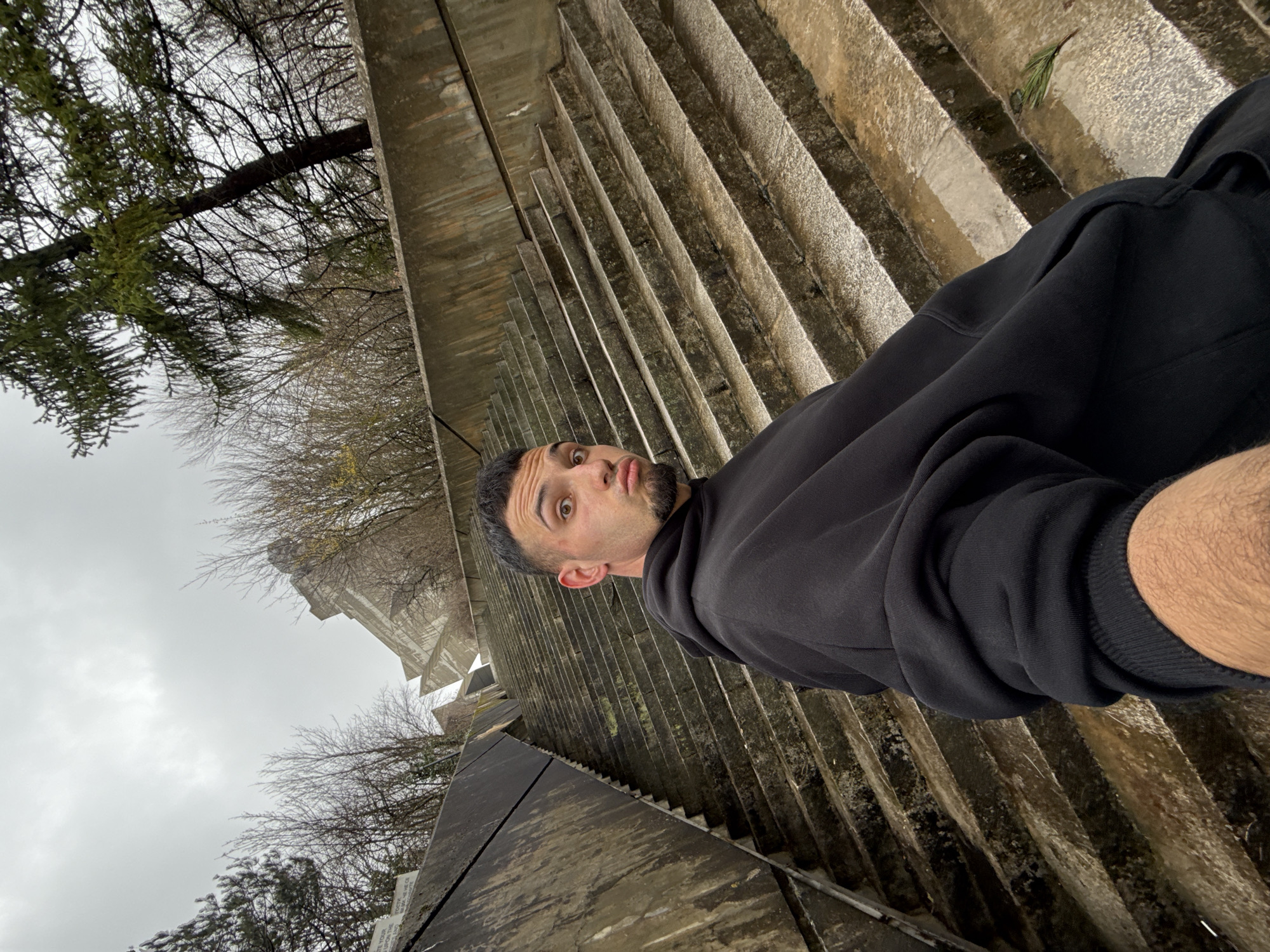 A man in a black hoodie stands on a concrete staircase complex, possibly the Monument to 1300 Years of Bulgaria in Shumen, Bulgaria. The sky is overcast. Trees are visible in the background. The man looks at the camera with an exaggerated expression. The staircase is weathered, with visible signs of age and moisture.