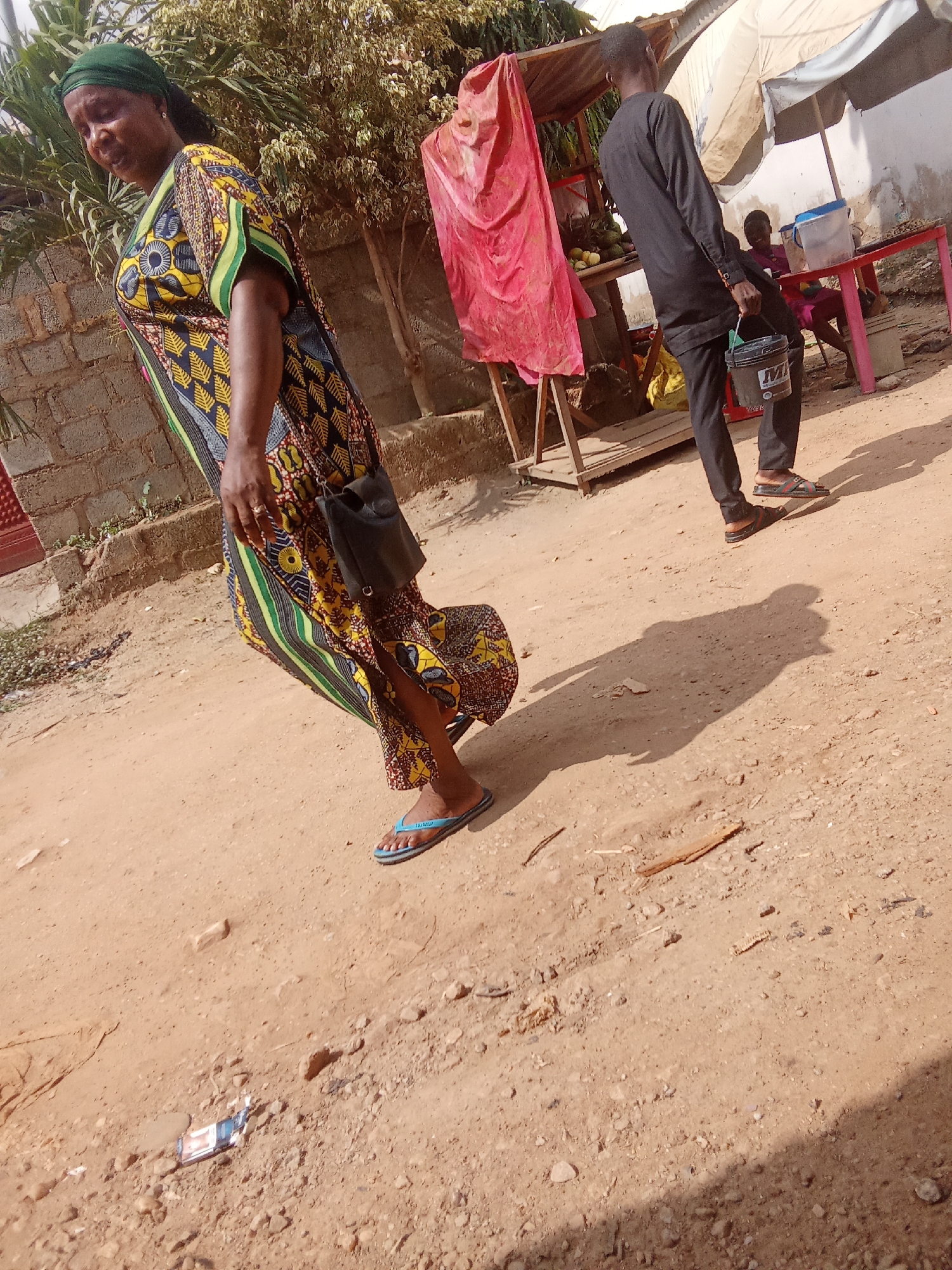 In Kagini, Nigeria, a woman walks in the foreground of a dirt path, her patterned dress flowing around her. She wears a green headscarf and blue flip-flops, and a small black bag is slung over her shoulder. Behind her, a man in a black shirt and pants walks away from the camera, carrying a black bucket. In the background, a market stall is set up with a red table displaying some items and a blue container. A woman is seated at the table, and a large pink cloth hangs from the stall. The scene appears to be in daylight, with the sun casting shadows on the ground. The overall impression is of a bustling street scene in a Nigerian town.
Visible text: On the black bucket the man is carrying, the letters "M" and "N" are visible.