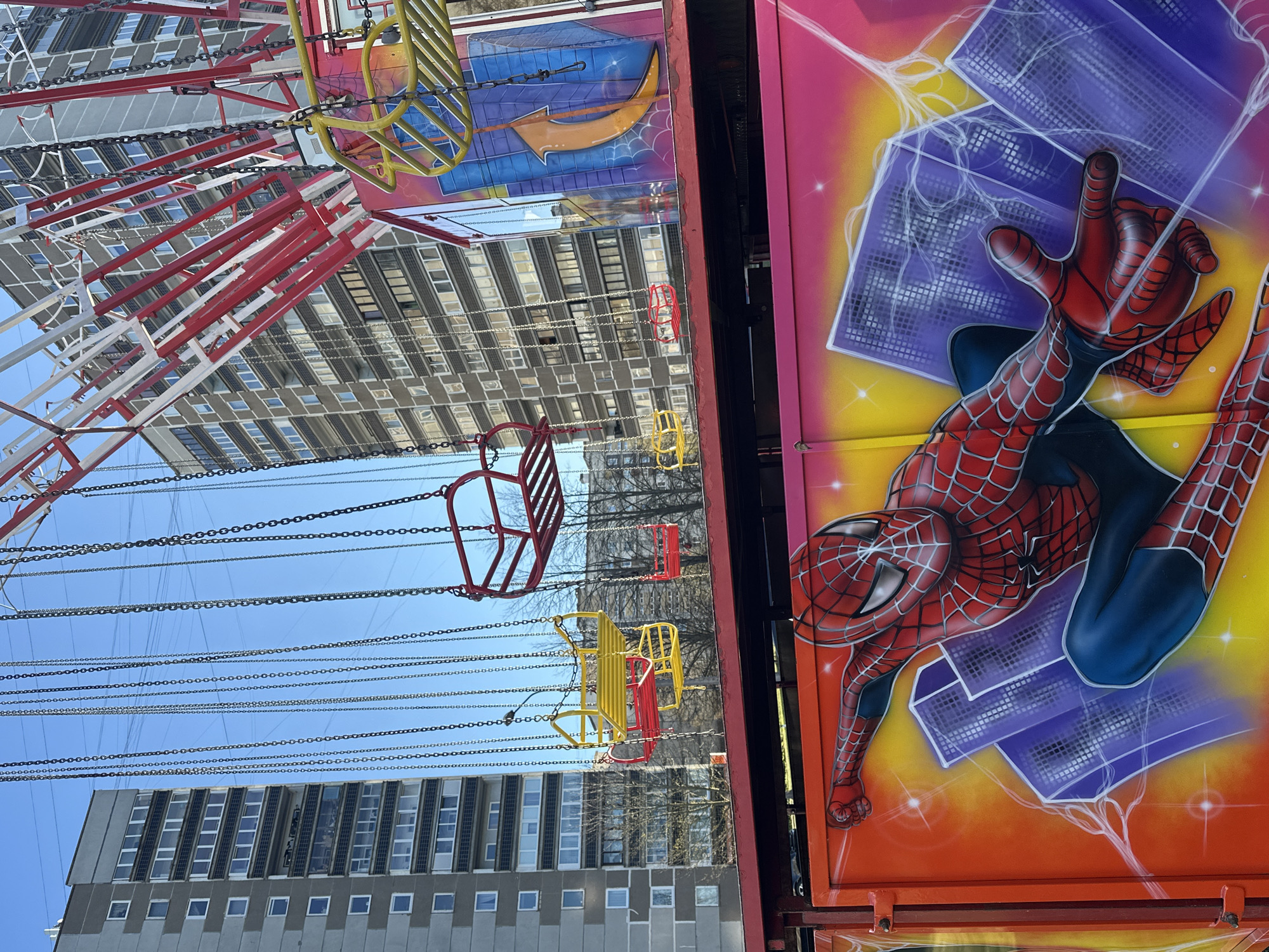 An outdoor scene captures a portion of a swing carousel ride set against an urban background under a clear blue sky. The image features a large, vertically oriented panel on the right side, vibrantly decorated with an airbrushed illustration of Marvel's Spider-Man. The superhero, in his classic red and blue suit, is depicted in mid-action, extending an arm as if shooting a web, with bright white web effects and glowing purple, checkered backgrounds against a striking gradient of pink, yellow, and orange.
To the left and extending into the background, the structure of the swing carousel is visible. Numerous empty swing seats, primarily colored red and yellow, hang from long chains, indicating the ride is not currently in operation. Behind the ride, several tall, modern buildings with repetitive window patterns, likely apartment blocks or offices, stand under the bright sky. The presence of bare tree branches between the ride and the buildings suggests it's a colder season, possibly late winter or early spring. The scene is illuminated by bright daylight, implying a clear, sunny day. The setup suggests a fairground or amusement park located within a city, possibly in Riga, Latvia. No discernible text is visible in the image.