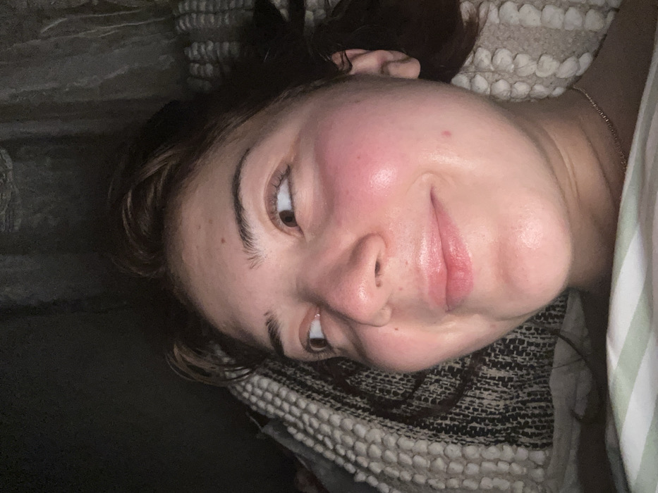 A young woman lies on a pillow, looking upwards with a slight smile. Her face is slightly flushed, particularly on her cheeks. She has brown eyes, full lips, and visible eyebrows. Her hair is dark brown, partially tied up. She's wearing a thin chain necklace. The pillow beneath her is patterned with a textured weave. Beside her is a dark, shadowy area.
