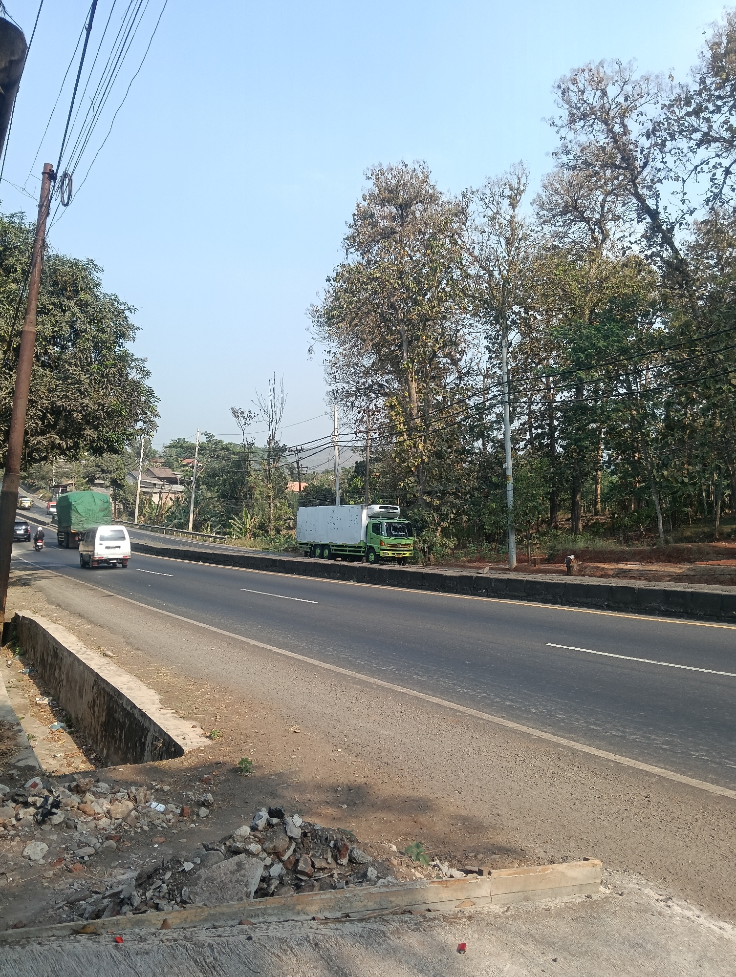 This is an eye-level, medium shot of a road in Adinuso, Indonesia, under a clear sky. The road is asphalt and divided by white lines. On the left side of the road, there is a concrete drain and debris. A utility pole with numerous wires stands prominently on the left. Trees line the right side of the road, with some appearing to be deciduous and bare. Several vehicles are visible on the road, including a white van, a green truck with a tarped cargo, and another white truck with a green cab. In the background, houses and more trees can be seen. The overall scene depicts a rural or suburban road environment.