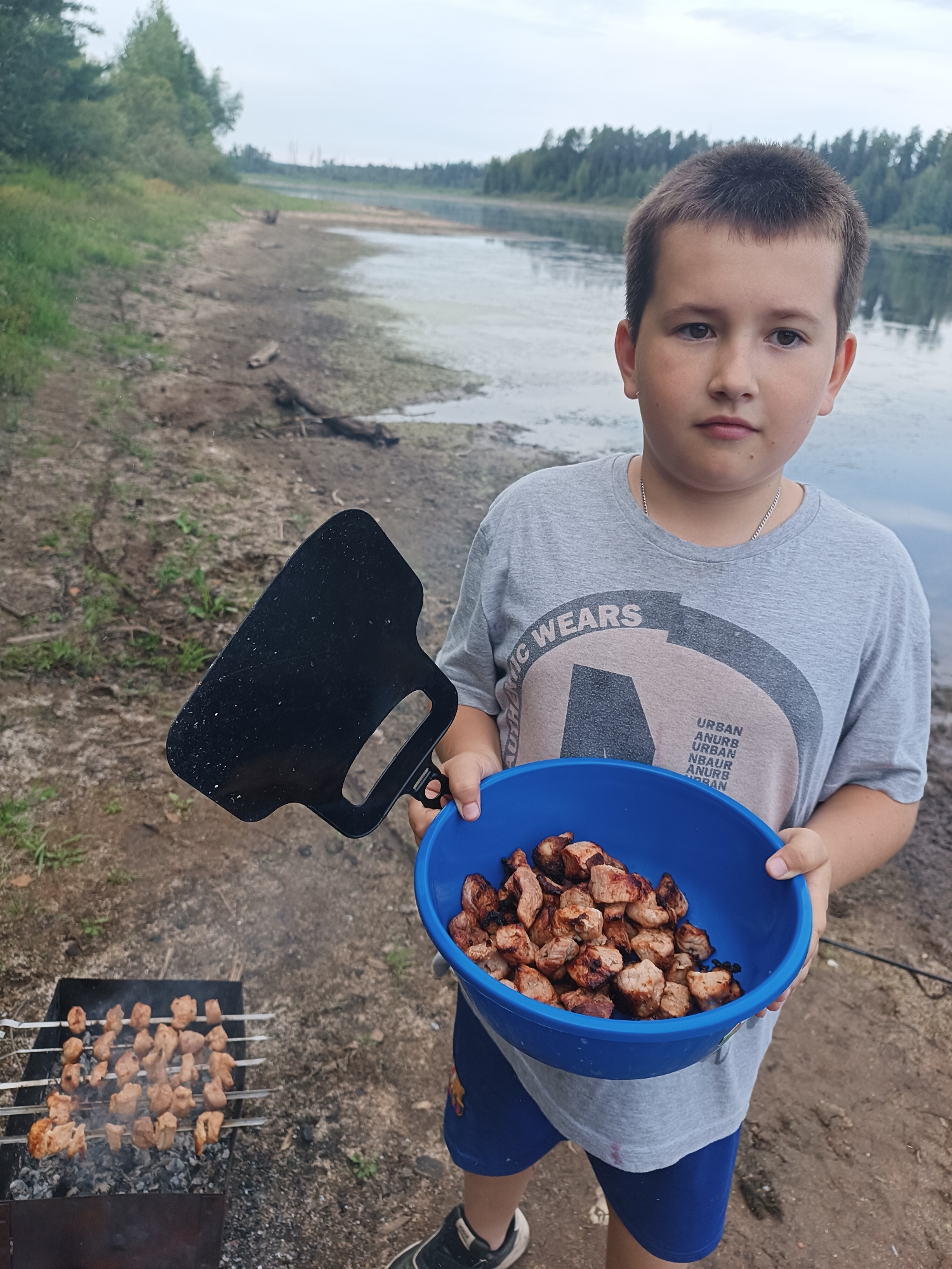 A young boy stands on the sandy shore of a lake in Opary, Russia, holding a blue bowl filled with cooked meat in his left hand and a black spatula in his right. He is wearing a grey t-shirt with a circular graphic and blue shorts. In the background, skewers of meat are grilling over a barbecue pit. The lake stretches into the distance, bordered by trees and a cloudy sky.