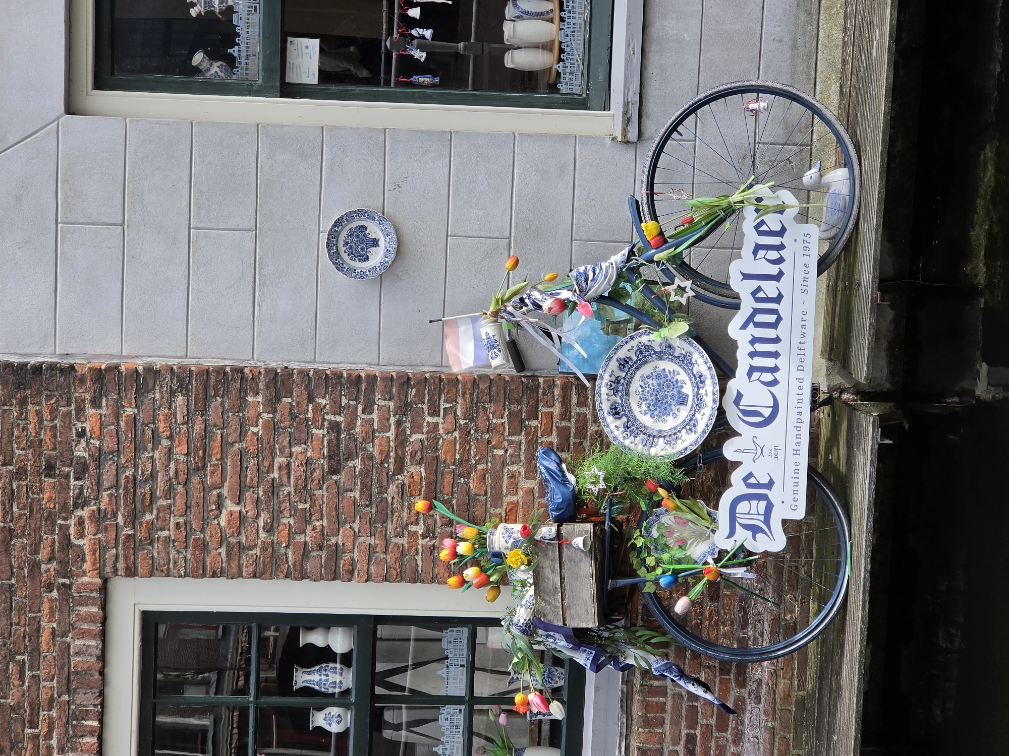 This image depicts a charming scene in the city of Delft, Netherlands, likely a shop front. A vintage bicycle, decorated with blue and white Delftware plates and colorful tulips, is parked on a wooden walkway. The bicycle's frame is adorned with floral arrangements and ribbons. One large Delftware plate is mounted on the wall behind the bicycle, and another is attached to the rear wheel. A sign on the bicycle reads "De Candelaer Genuine Handpainted Delftware Since 1975," indicating the presence of a Delftware shop. The background features a brick building on the left with a window displaying Delftware vases, and a light gray stone building on the right with a window showcasing more Delftware items. The setting appears to be a canal or waterway, as suggested by the dark water visible below the walkway.