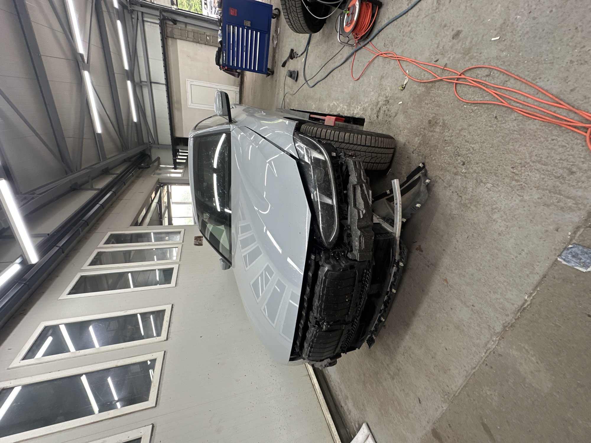 An interior shot captures a light grey car with significant front-end damage parked in what appears to be a workshop or garage. The car is positioned at an angle, with its front facing towards the right side of the frame. The entire front bumper and grille assembly are missing or severely broken, exposing the underlying structural components and mechanics of the vehicle. Reflections of overhead fluorescent lights are clearly visible on the car's hood and side panels.
The setting is an industrial-style space with a high ceiling supported by metal beams. Numerous bright, rectangular fluorescent light fixtures illuminate the area, running in rows along the ceiling and walls. The floor is made of concrete, showing various scuffs, stains, and a visible seam running across it.
In the background and around the car, various workshop items are scattered. To the left, a blue, multi-drawer toolbox on wheels is visible. Nearby, spare tires and an orange extension cord coiled around a reel sit on the floor. Another segment of the orange extension cord snakes across the concrete floor towards the lower right. A black air hose or cable is also present.
A large garage door with multiple rectangular windows, angled diagonally, occupies the left side of the frame, reflecting the interior lights. Another plain white door is visible in the distance. The scene suggests a vehicle undergoing or awaiting repairs in a functional, well-lit garage environment. No people or visible text are present in the image.