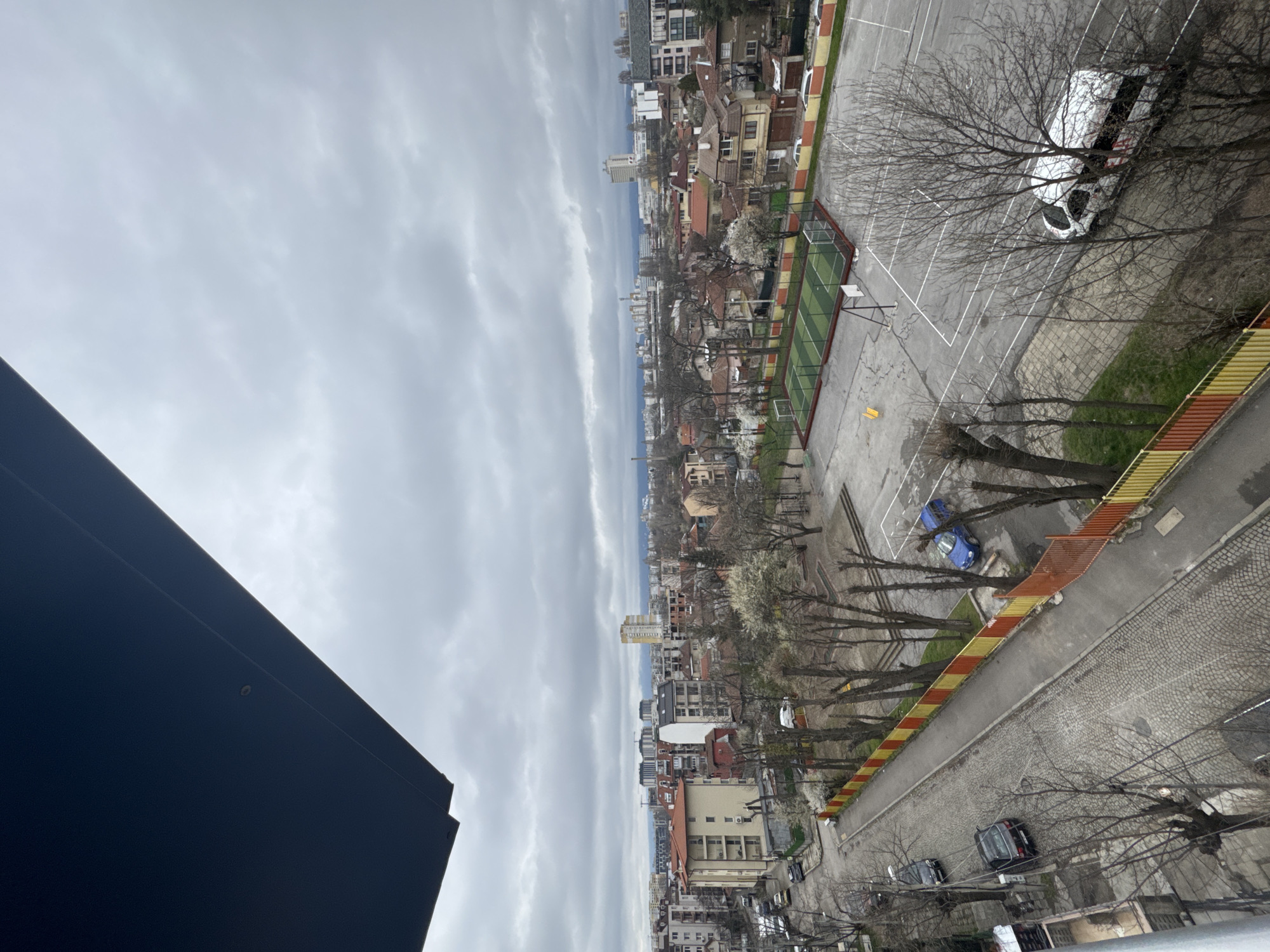 The image shows an elevated view of a cityscape, likely taken from a high-rise building in Sofia, Bulgaria. A portion of a modern building's dark-colored facade is visible in the foreground. The city itself consists of a mix of low-rise residential buildings with red-tiled roofs and a few taller, modern structures in the distance. A schoolyard with a green sports field is visible on the right side. A white van is parked on the schoolyard, surrounded by bare trees. Parallel to the schoolyard runs a street with a distinctive yellow and red striped barrier. A few parked cars are visible along the street. The sky is overcast and filled with gray clouds. The general atmosphere suggests a cloudy, possibly springtime day.