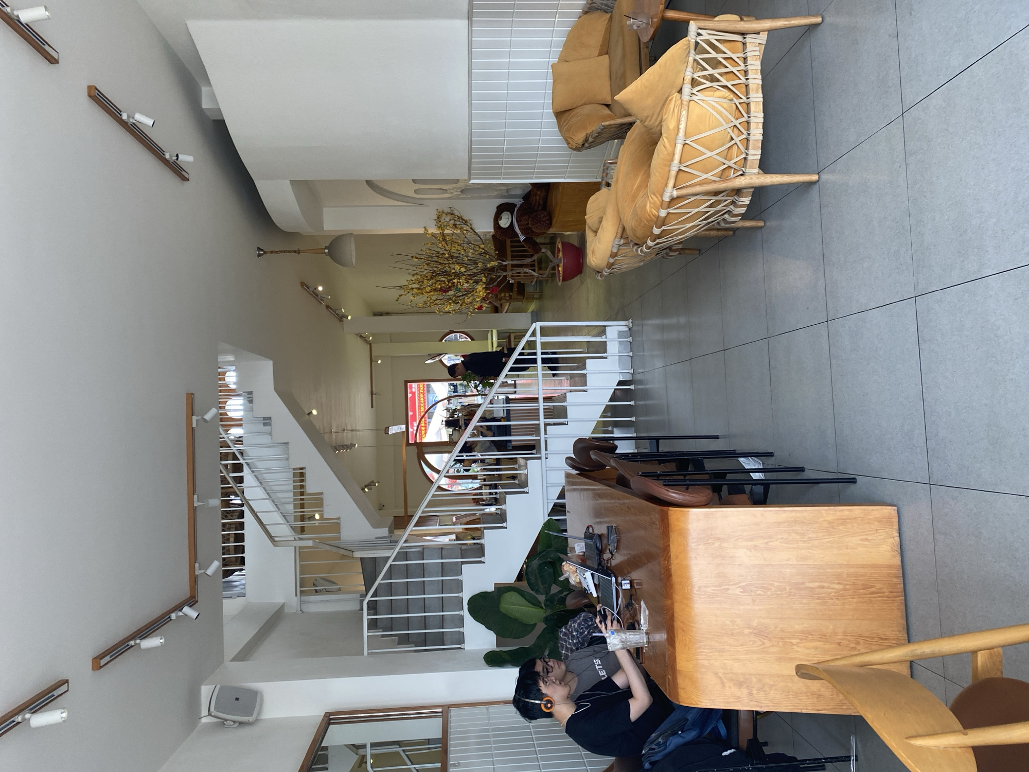 The image captures an interior view in Lâm Viên, Đà Lạt, Vietnam. The location features a multi-level space with a staircase lined with white railings. Seating arrangements include wicker chairs with gold cushions, situated on the upper floor. A long wooden table with chairs is visible, with a person wearing headphones and glasses working on a laptop. The environment includes lighting fixtures on the wall, a large potted plant, and architectural details of a modern, open space. The staircase leads to a lower level. The space appears to be a café or coworking area.