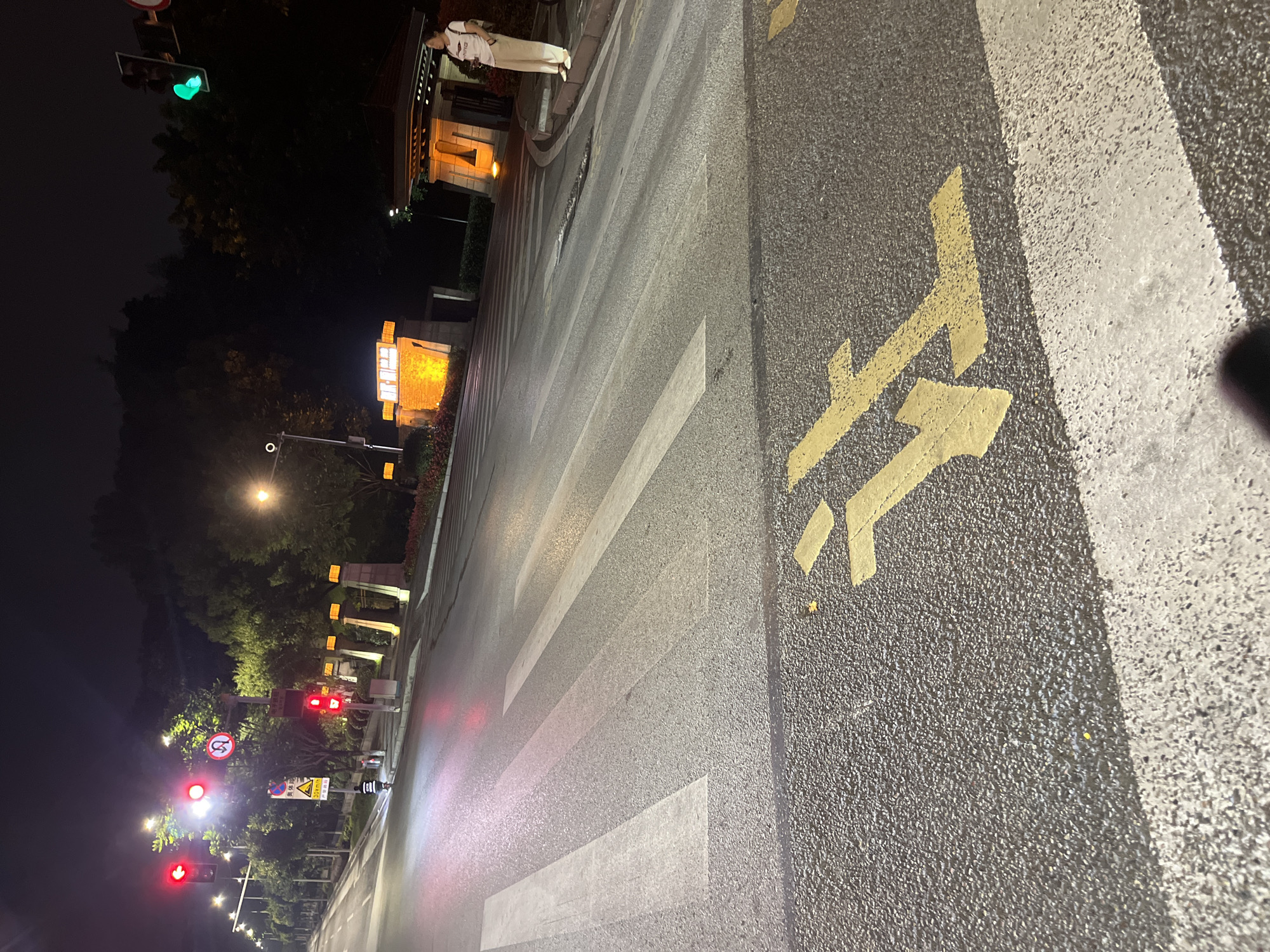 The image depicts a nighttime scene on a city street in Jiulongpo District, China. A pedestrian crossing is in the foreground, marked by white stripes on asphalt. Yellow directional arrows are painted on the road surface, indicating a right turn. Traffic lights are visible in the distance, with one showing a green light. A woman in light-colored pants and a white t-shirt stands on the sidewalk to the right, holding a camera. The street is illuminated by streetlights and the glow from buildings. Trees and foliage line the sides of the road, and a dark sky suggests it is late at night. The overall atmosphere is calm and urban.
