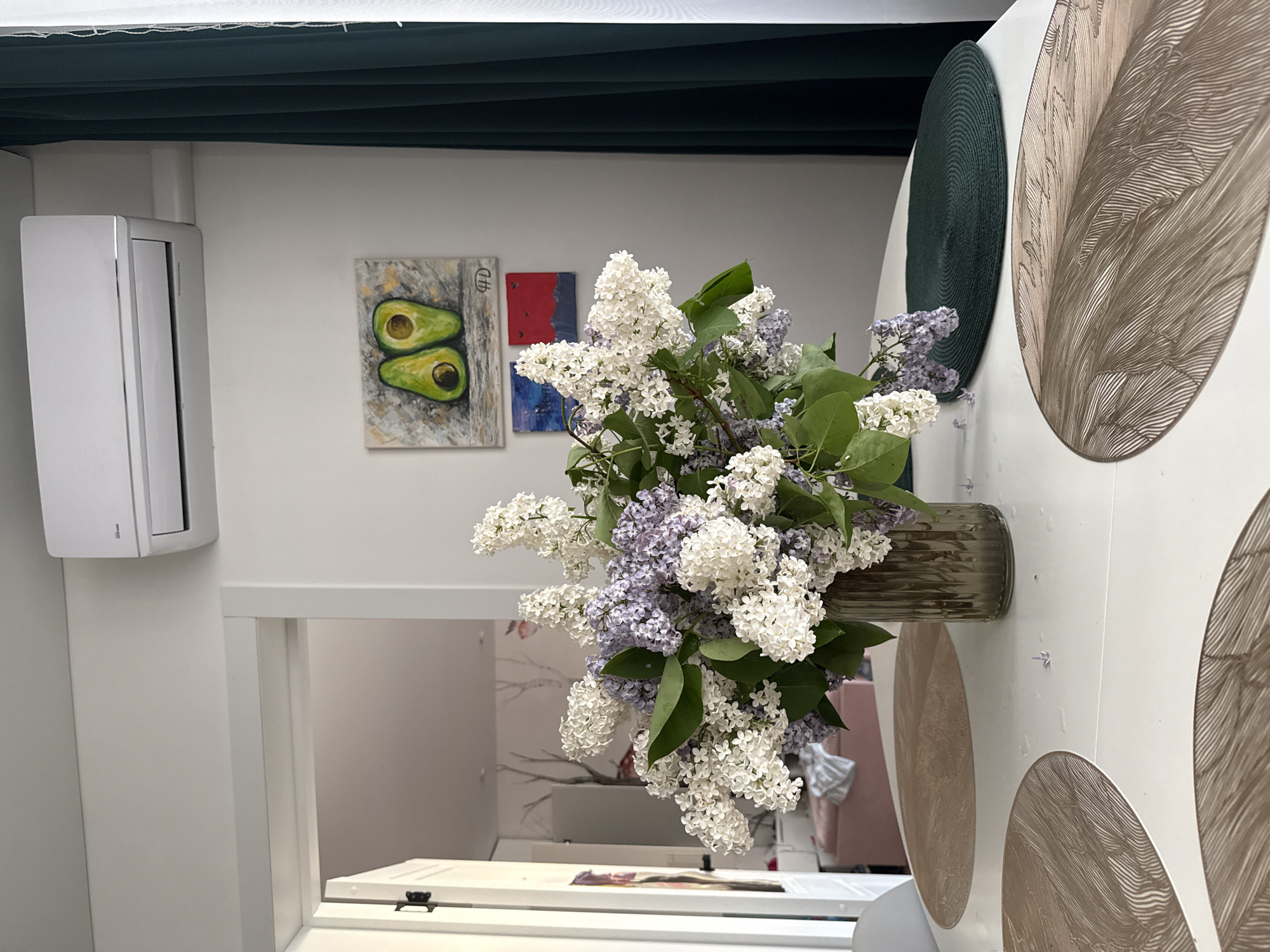 A slightly rotated medium shot depicts a bouquet of white and purple lilacs in a textured grey vase. The vase is positioned on a white surface adorned with large, round, decorative elements. One is a dark green woven disc, and the other two are light brown with etched patterns.
Behind the bouquet, mounted on the wall, are two small paintings: one with an image of two avocados and another with red and blue squares.
In the background, through an opening, is a room with a table and chairs, along with wallpaper featuring tree branches and birds. A pink chair is also visible in the room. A white air conditioning unit is mounted on the wall to the left of the opening.
The photograph was likely taken indoors. There is no indication of its location in the city of Veliky Novgorod, Russia.