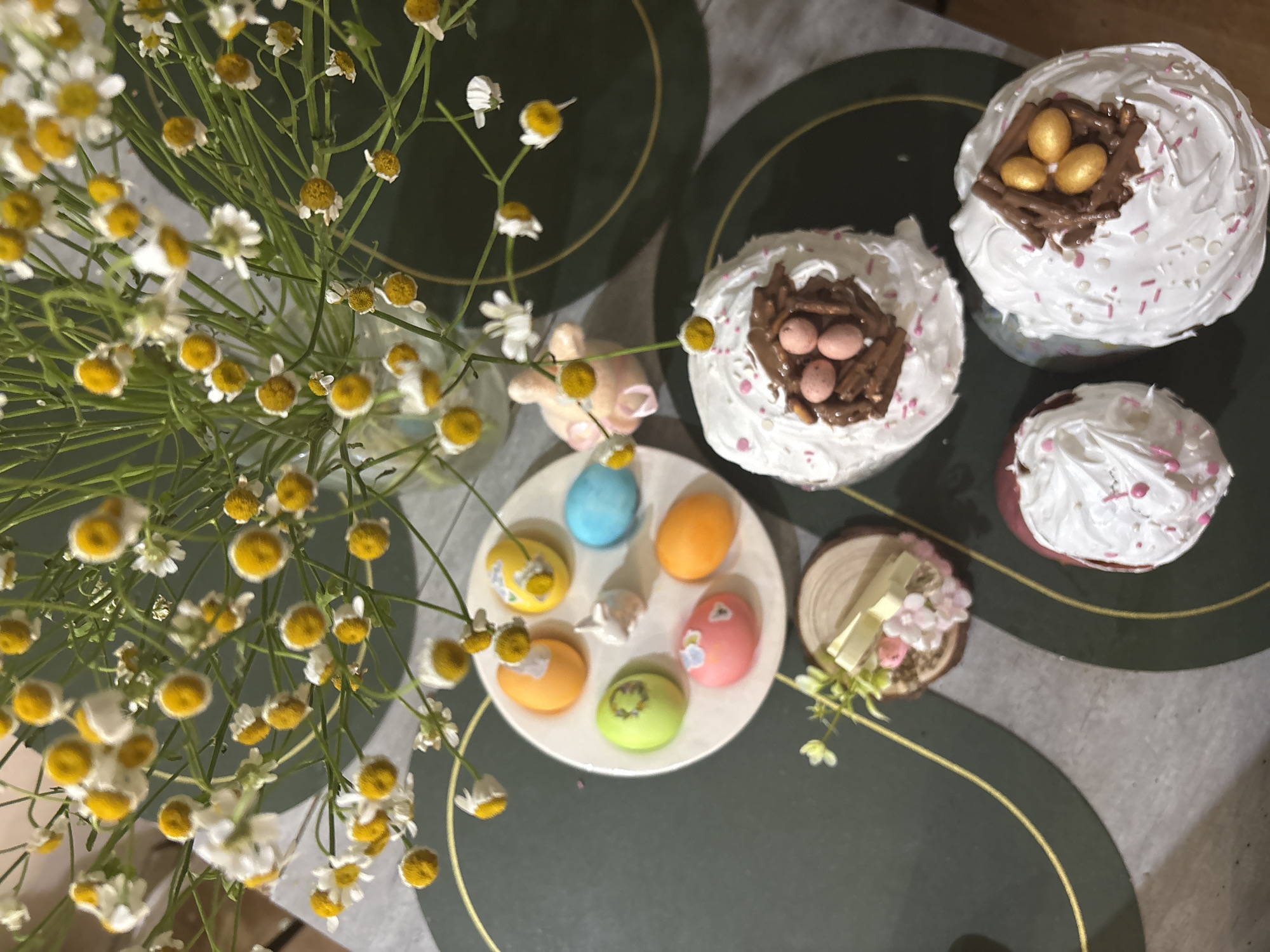 The image shows a still life of Easter treats and decorations. A bouquet of small white and yellow flowers is in the foreground, slightly obscuring a white plate containing several pastel-colored Easter eggs. Next to the plate is a small decorative figure of a piglet. Several Easter cakes with white frosting and chocolate "nests" filled with candy eggs are arranged on the right side of the image. A small wooden disk with flowers and ribbon is also present. The scene is set on a table with a dark green and light gray placemat with gold accents. There is no clear indication of the location being Chaikovsky, Russia.