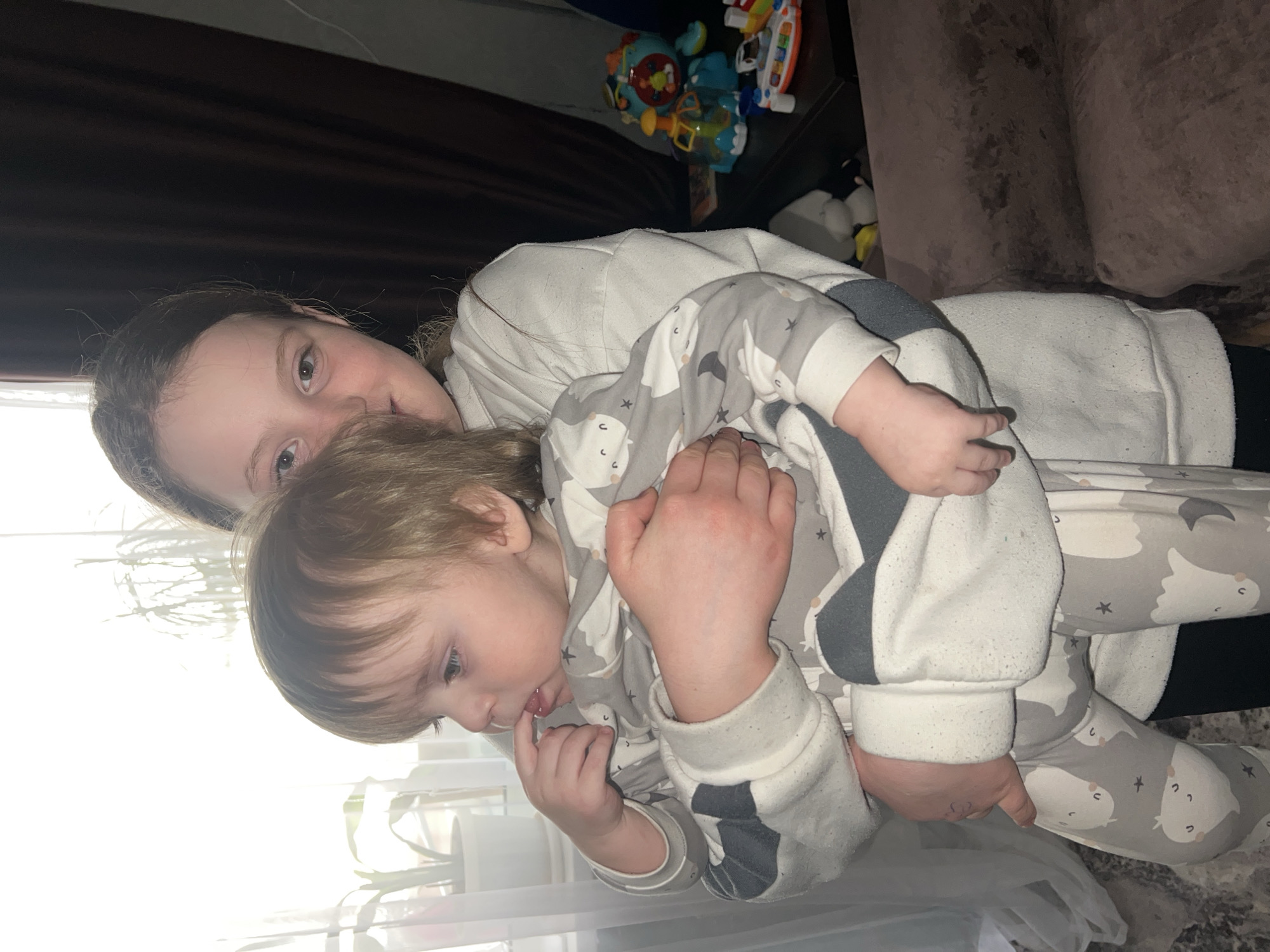 This image depicts a residential interior in the city of Noviy, Russia. A female adolescent with light brown hair, wearing a light-colored long-sleeved top, holds a toddler. She is positioned in the center-right, looking towards the camera with a slight smile. The toddler, with blonde hair, is dressed in a grey and white jumpsuit patterned with cartoon ghosts and stars, and is looking to the left, with a finger in their mouth. The background features a brightly lit window with white sheer curtains and a potted plant on the left. To the right, dark brown curtains and a dark table are visible, upon which several colorful children's toys are arranged, including a blue and orange electronic toy. A brown sofa is partially visible in the lower right, and a mottled floor surface is discernable in the lower left.