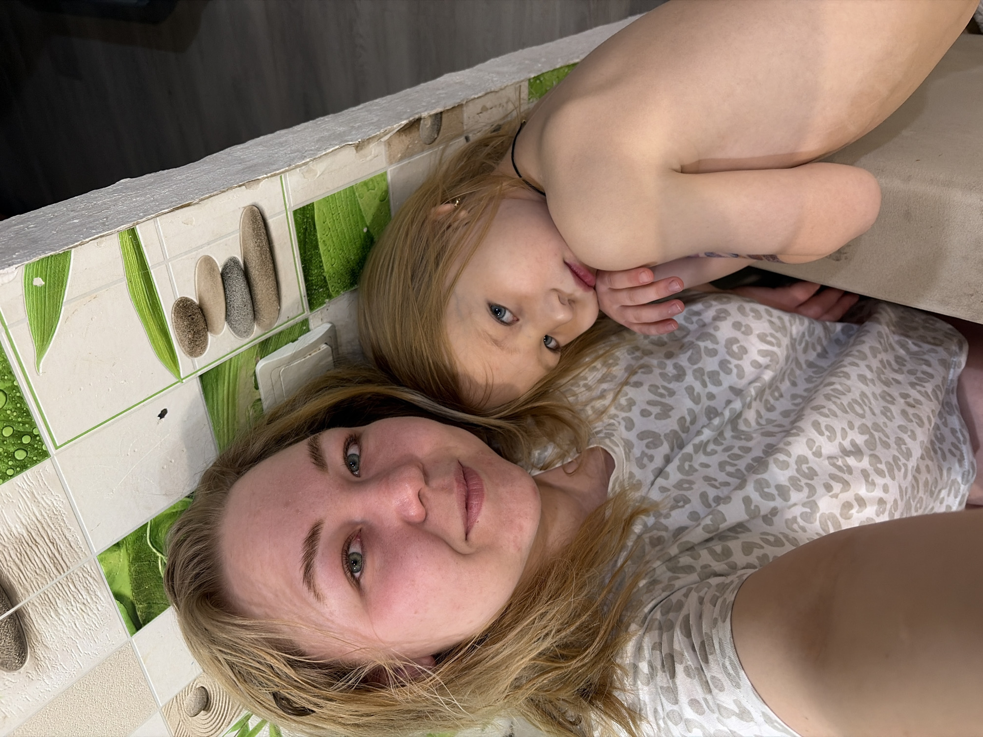 The image shows a woman and a young child lying down. The woman has fair skin, blonde hair, and green eyes. She is wearing a gray and white patterned shirt. The child has fair skin, blonde hair, and blue eyes. The child is lying with their hand near their mouth. They are lying on a patterned surface next to a tiled wall with green and pebble designs. The location is likely a domestic interior, potentially in Noviy, Russia.