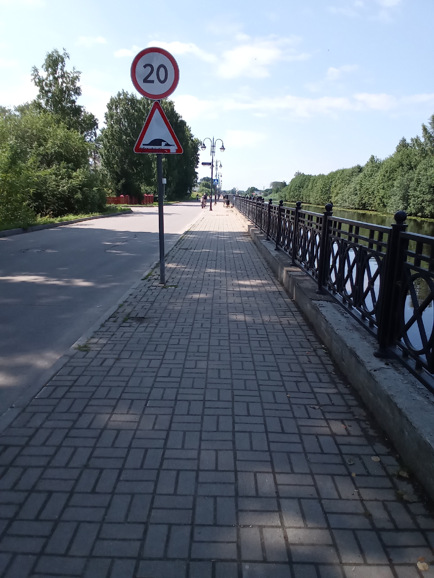 The image depicts a paved pedestrian walkway next to a road, with a body of water bordered by trees to the right. A black metal fence separates the walkway from the water. Two traffic signs are visible: a circular sign with "20" in black on a white background with a red border, indicating a speed limit of 20, and a triangular sign below it with a red border and white background, depicting a speed bump. Streetlights line the walkway, and in the distance, a few people are walking or cycling. The sky is blue with scattered clouds. This scene is located on the embankment of the Beloye Lake in the city of Belozersk, Russia.