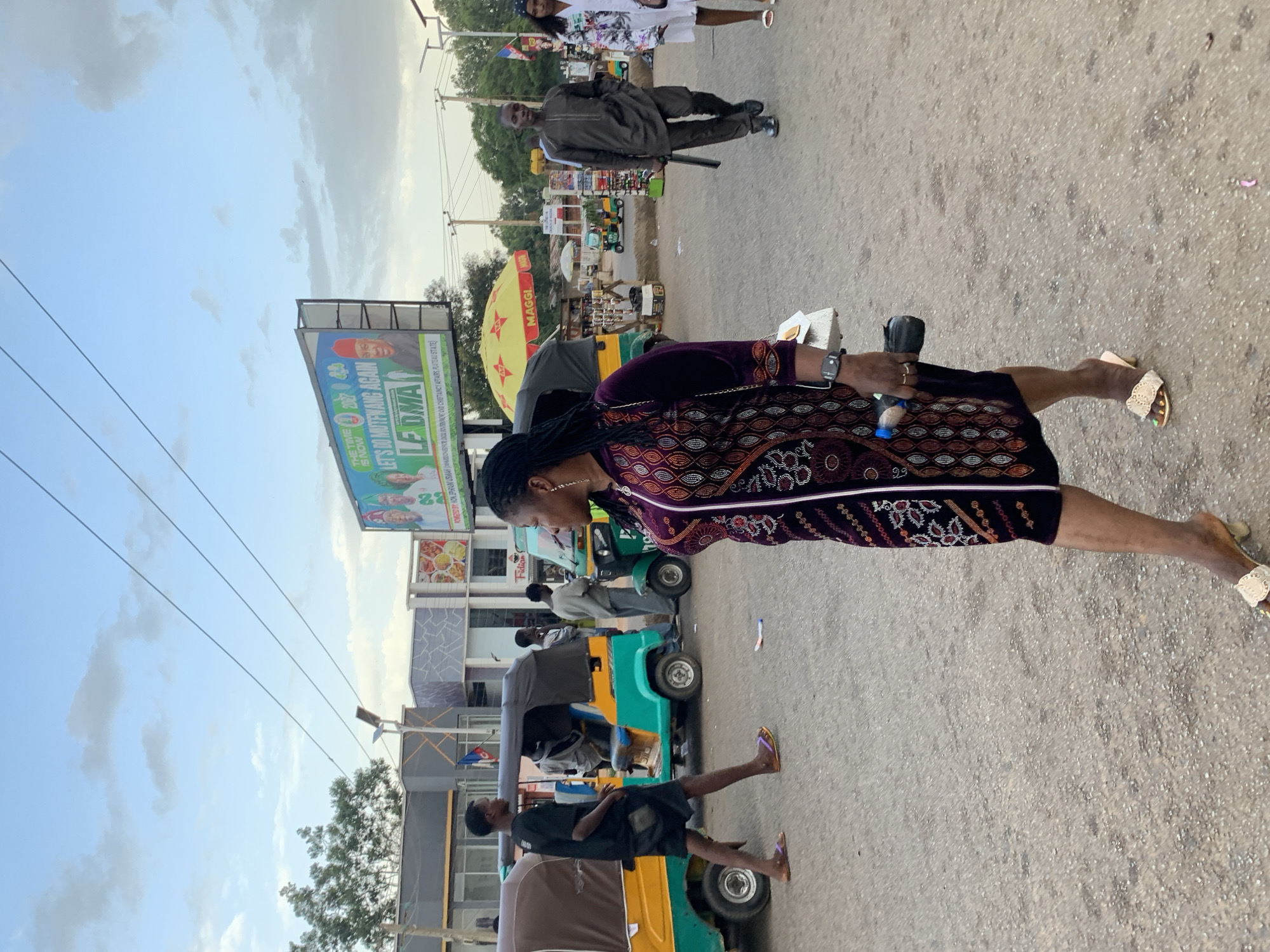 The image depicts a street scene in Jos, Nigeria, likely a market area or a busy intersection. The sky is cloudy with hints of blue, and overhead power lines are visible. In the background, a large billboard displays political campaign imagery with text including "THE TIME IS NOW" and "LET'S DO MUTFWANG AGAIN". Several people are present, including a woman in the foreground wearing a patterned dress and high heels, carrying a water bottle and a purse. To her left, a young man in a black t-shirt and shorts walks past a bright yellow and teal auto-rickshaw. Further down the street, another auto-rickshaw is parked, and several other individuals are seen, some interacting with street vendors under a yellow umbrella bearing the "MAGGI" logo. The ground is paved with asphalt, showing signs of wear and tear. Buildings with modern architecture line the street, some featuring patterned facades. The overall atmosphere suggests a bustling urban environment in Jos.