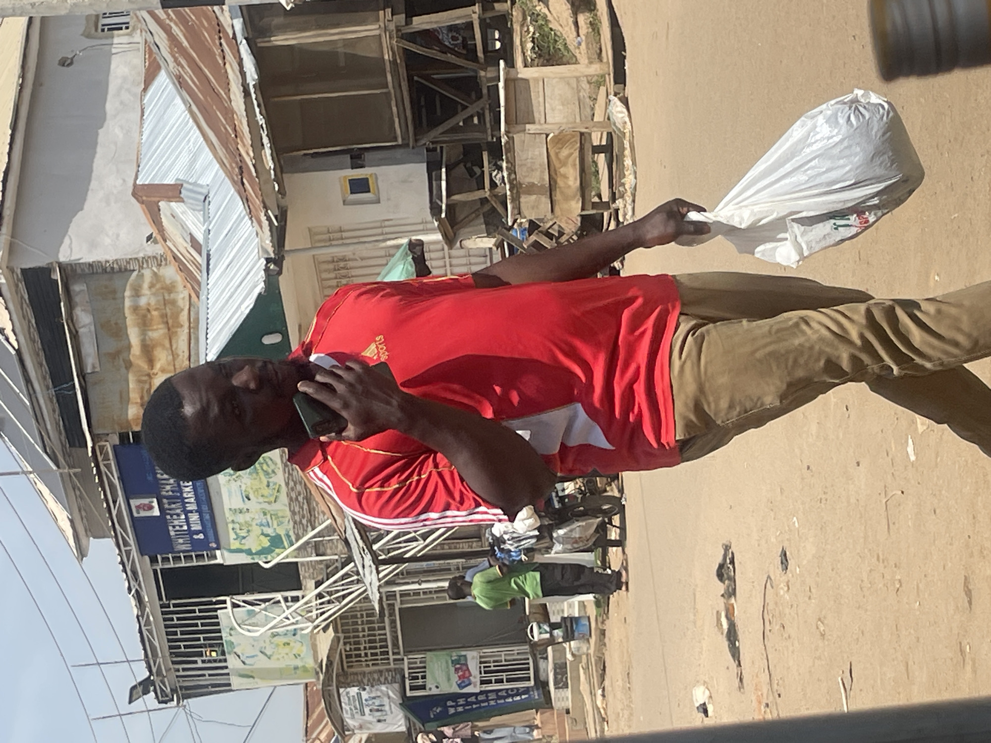 A Black man in a red t-shirt and tan pants walks down a dusty street in Abuja, Nigeria. He holds a cell phone to his ear with his right hand and a white plastic bag with his left. Behind him, a building with a blue sign reads "WHITEHEART PHARM & MINI-MARKET". Other structures with corrugated metal roofs line the street, and debris is scattered on the ground.