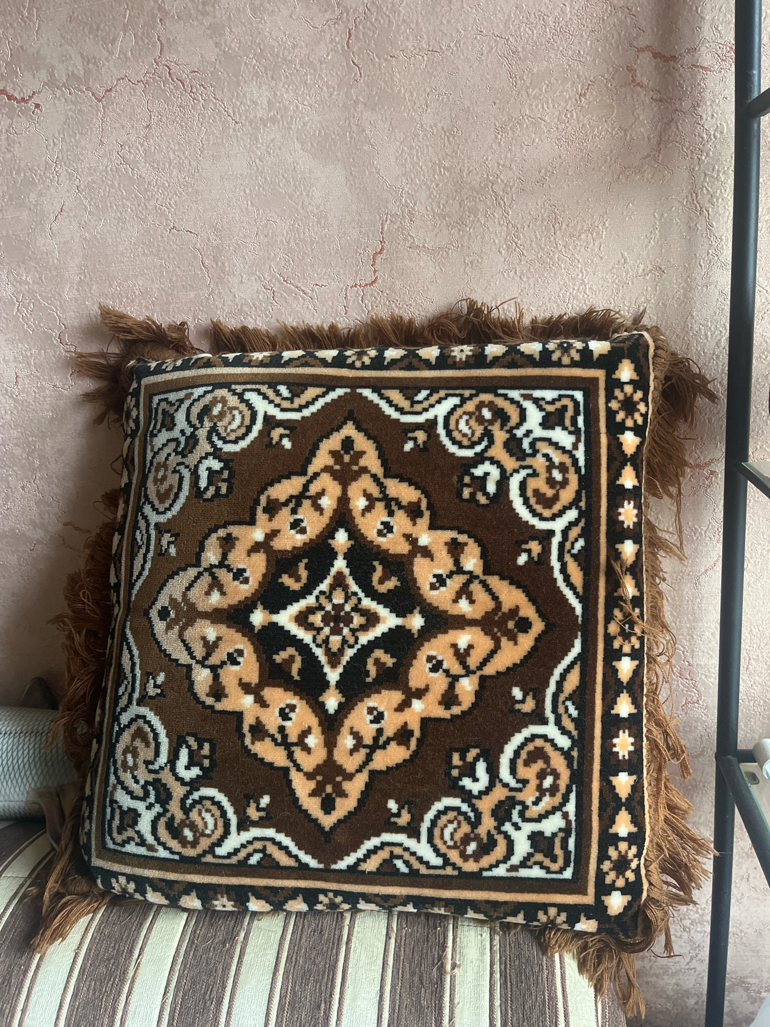 A close-up shot shows a brown throw pillow with a geometric pattern, fringed on all sides, resting on a striped surface. The pillow's intricate design features shades of brown, tan, and white, with a central medallion and floral motifs. The background is a textured, pale pink wall with subtle cracks and a dark metal object on the right. The setting appears to be indoors, possibly a home in Vladimirovka, Russia. There are no people or animals visible in the image.