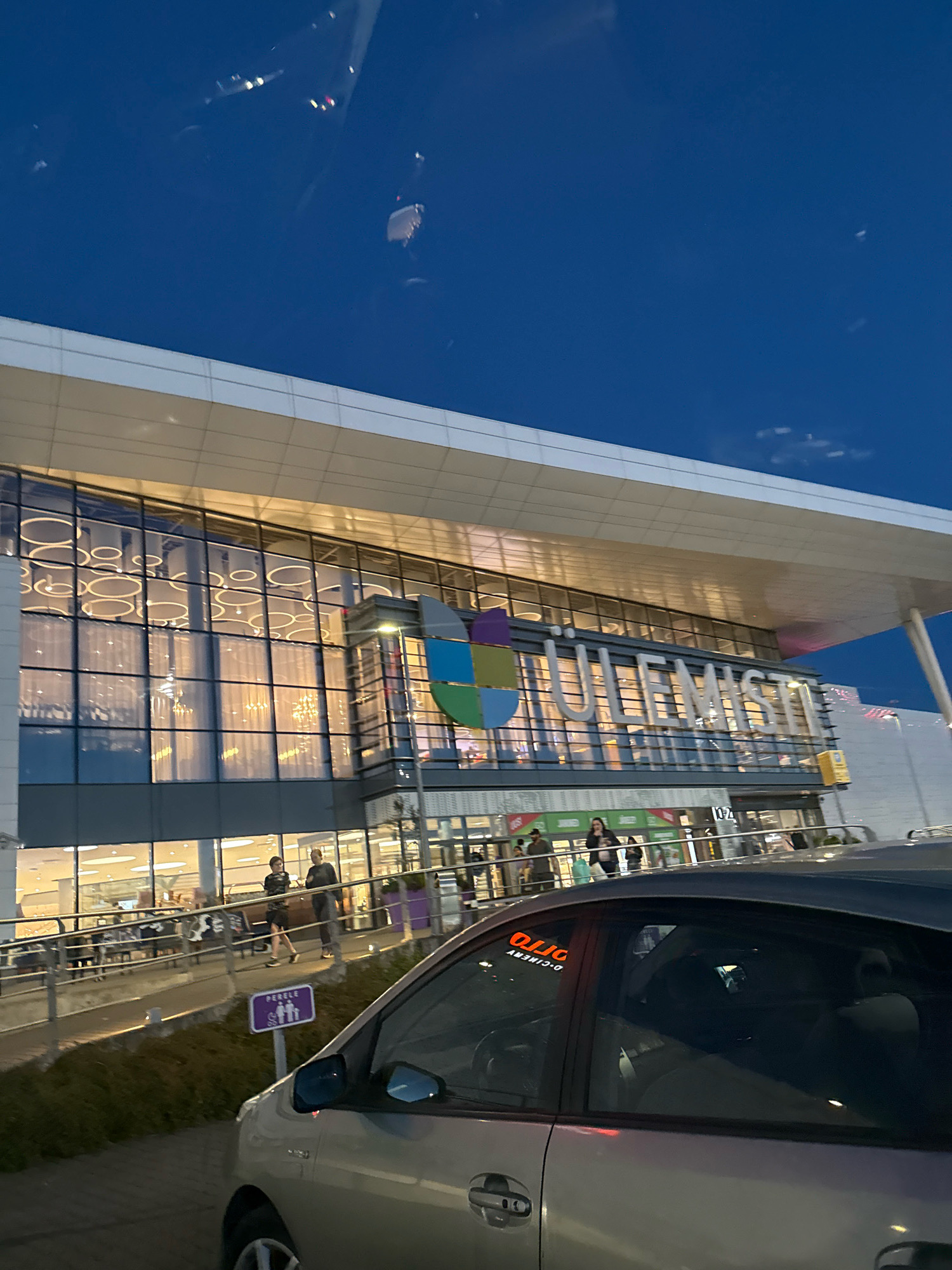 The image shows the Ülemiste Keskus shopping mall in Tallinn, Estonia, at dusk. The mall's facade features large glass windows reflecting the interior lights and a prominent sign with the mall's name. A silver car is parked in the foreground, obscuring the lower part of the building. A sign with a purple background and white figures representing a family is visible near the car, indicating a family parking spot. People are seen walking near the entrance of the mall. The sky above is a deep blue, with some clouds and reflections of lights.