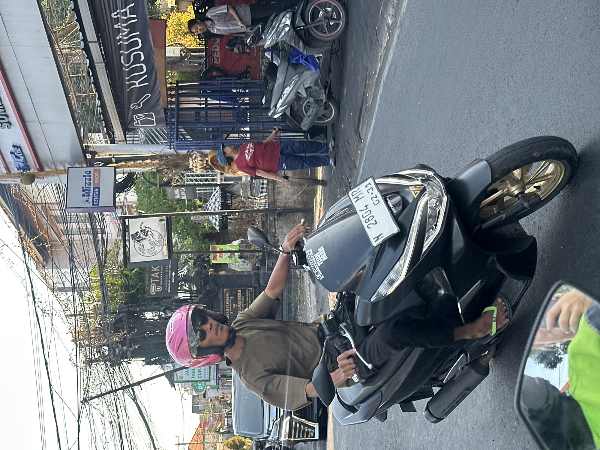 This is a street-level, eye-level, eye-level photograph taken on a street in North Kuta, Indonesia, during daylight hours with bright sunlight creating strong shadows. The primary subject is a person riding a black scooter. The rider is wearing a bright pink helmet and sunglasses, a dark olive-colored t-shirt, and black pants. Their right hand is on the scooter's handlebar, and their left foot, clad in a lime green flip-flop, is resting on the scooter's footboard. The scooter is a modern, black model with gold accents on the wheels. A license plate reading "N 2804 MR 02-31" is visible on the scooter.
In the background, a dark-colored car is parked on the left side of the street. On the right side of the street, there is a sidewalk with several people and commercial signage. One woman, wearing a red shirt and blue pants, is sweeping the sidewalk with a broom. Another woman, wearing a grey uniform, stands near a blue gate. Behind them, banners display text including "KUSUMA" and a logo with tools. Buildings with multiple stories and abundant overhead electrical wires are visible. Signs for businesses such as "Mizzle Center Shop" and "TAK COFFEE & EAT" are also present. The street surface is asphalt. The overall scene depicts a typical urban street environment in Indonesia, likely a commercial area.