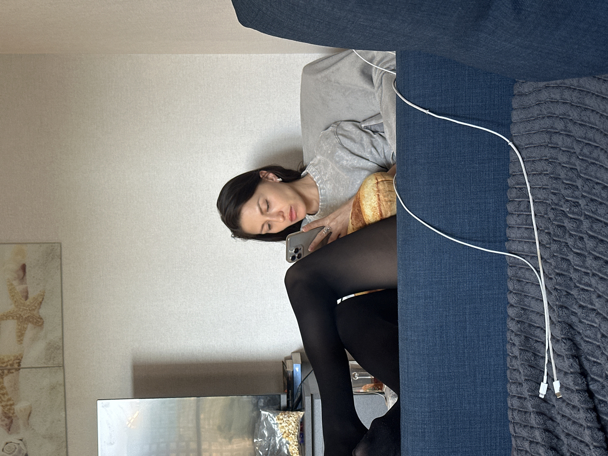 The image, rotated 90 degrees clockwise from its intended orientation, depicts a woman relaxing on a sofa in an indoor setting, likely a living room in Murino, Russia. The woman has long dark hair and is wearing a light grey sweatshirt and black opaque tights. She is seen from the waist up, reclining on a blue fabric sofa, with her eyes closed or partially closed, suggesting a moment of rest or contemplation. She holds a silver smartphone with a visible multi-lens camera setup in her hands, and a distinctive ring is visible on her right hand.
Next to her on the sofa is a whimsical cushion designed to resemble a loaf of bread. A white charging cable is draped over the armrest of the sofa, which is also partially covered by a textured grey blanket. In the background, light-colored walls are adorned with two framed pictures featuring beach-themed motifs, including starfish and seashells. Below these decorations, the top portion of a television screen or monitor is visible, and to its right, a shelf holds books and a clear bag containing what appears to be popcorn or similar puffed snacks. The overall scene conveys a calm and relaxed atmosphere. No explicit text is visible in the image.