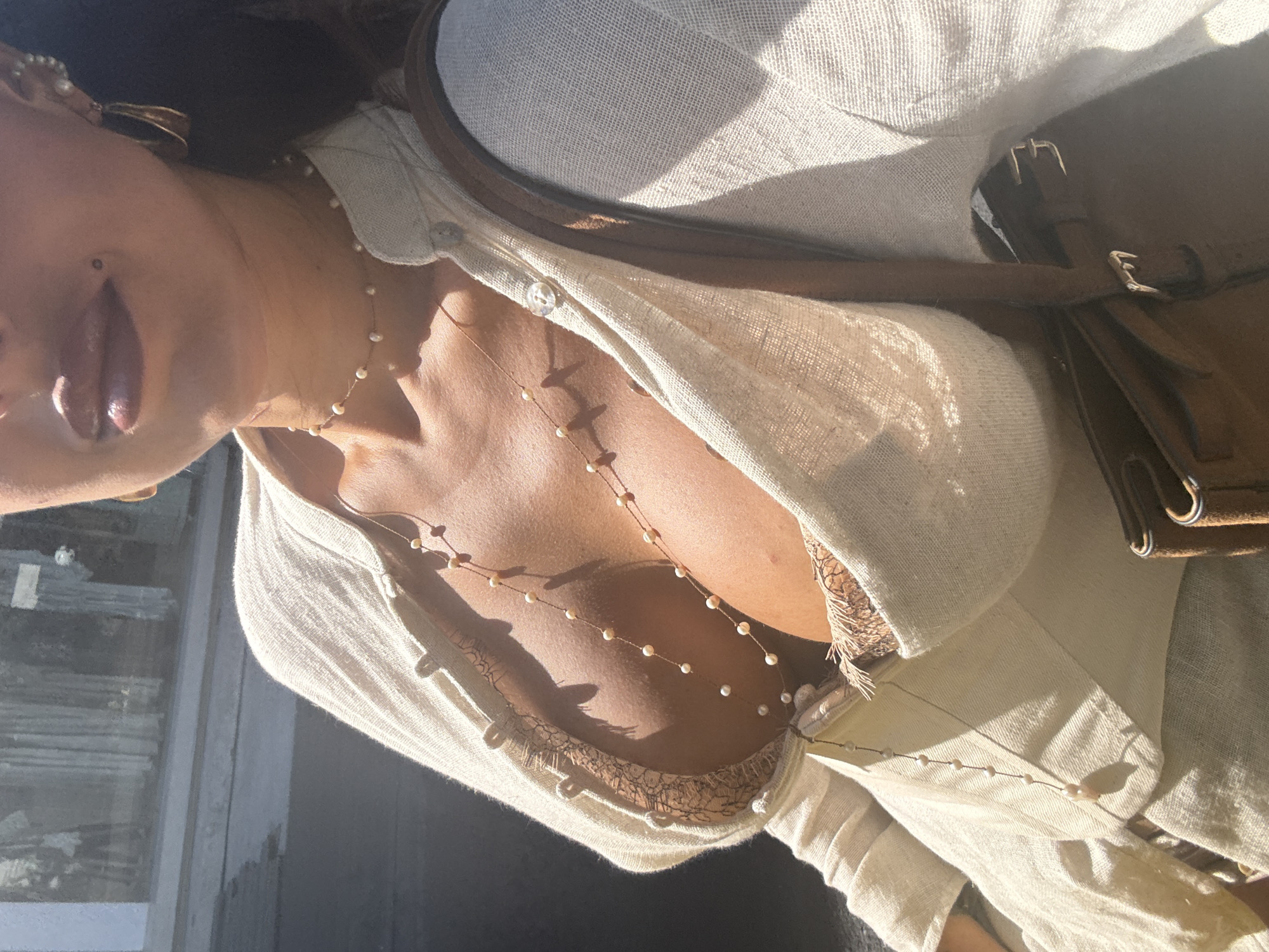 A medium shot of a woman's chest and shoulders wearing a light-colored button-down shirt and two pearl necklaces. The shirt is partially unbuttoned, revealing a lace-trimmed bra. A brown leather bag with a silver buckle is visible over her shoulder. Her face is partially visible, showing a lip piercing. The photo is taken in daylight, possibly outdoors. No information indicates a location in North Kuta, Indonesia.