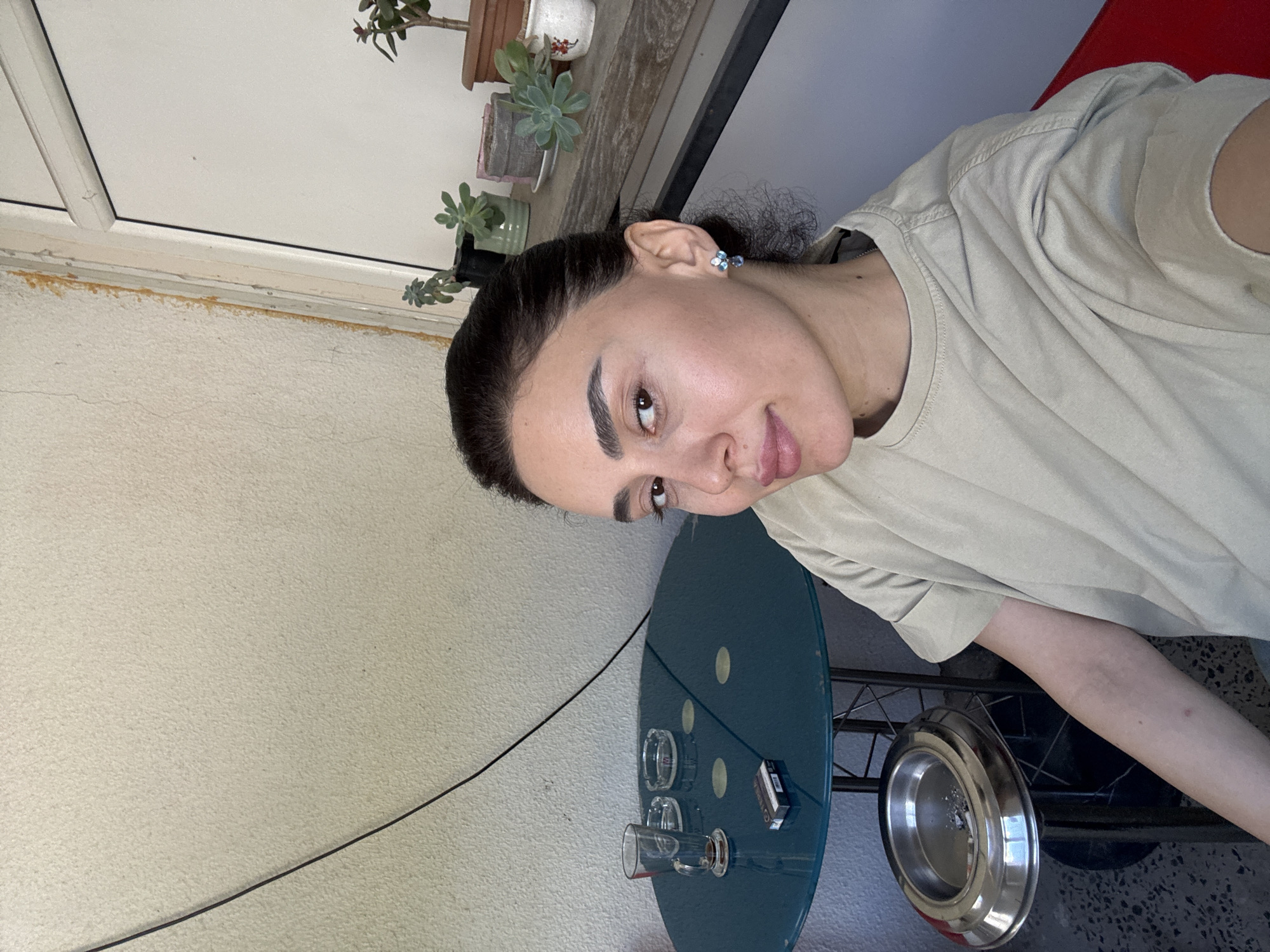 The image shows a person with dark hair and light skin wearing a beige t-shirt. They are standing on a balcony, evidenced by the presence of potted succulents along a window ledge. The balcony's wall is painted off-white. A small blue round table sits on the balcony, holding glassware, an ashtray, and a pack of cigarettes. Based on the style of architecture and balcony, the location may be in Tbilisi, Georgia. The person is looking directly at the camera. There is a wire cable snaking down the wall.