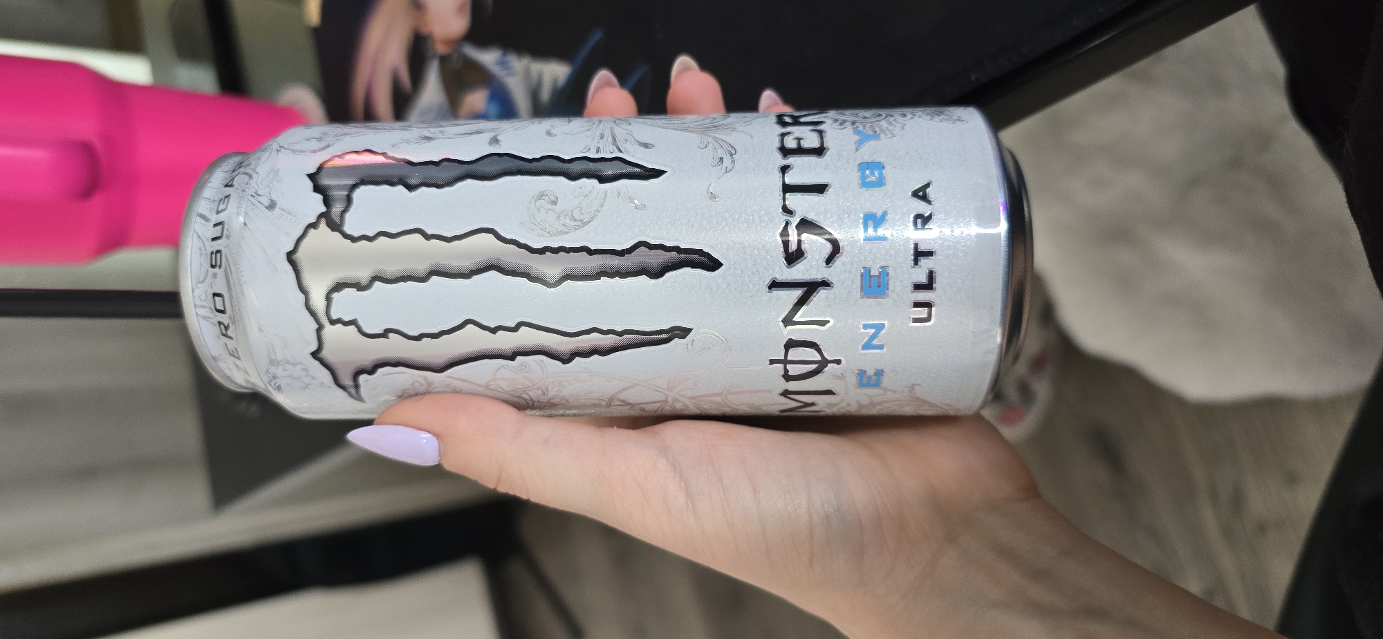 A person's hand holds a can of Monster Energy Ultra drink. The can is white with a silver Monster logo. The thumb of the hand has a lavender nail. The background is blurry, but a pink object and a framed photo are visible. The location is unknown, not specified. The image does not contain evidence that this photo was taken in the city of Burgas, Bulgaria.