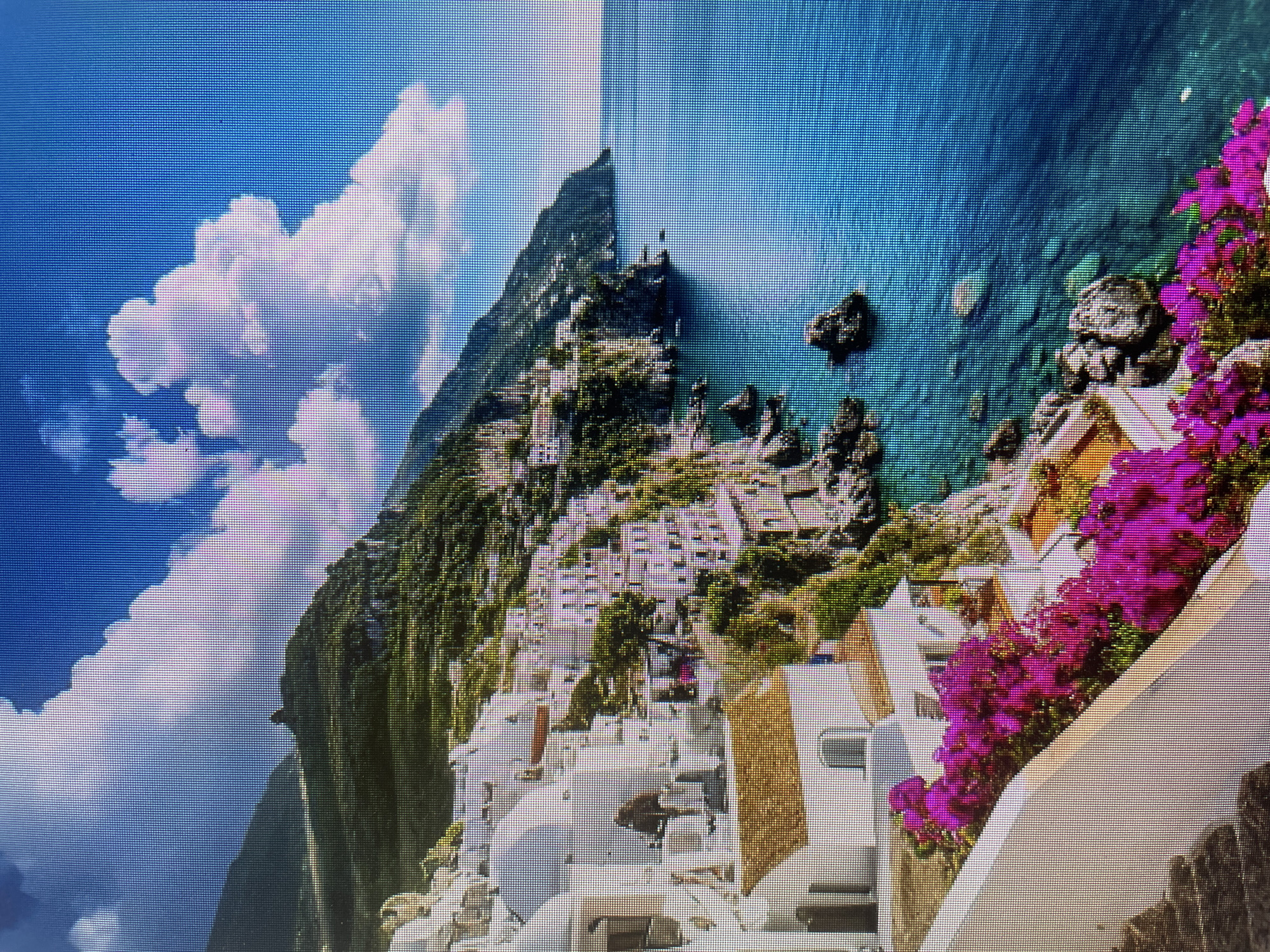 This media file captures a picturesque, vibrant coastal town built into a steep, verdant hillside overlooking a tranquil blue sea. The image itself appears to be a photograph of a digital screen, as indicated by the prominent grid of pixels visible across the entire frame and a subtle vertical line down the center, suggesting a display artifact or seam.
The scene features numerous white-washed buildings with light-colored, possibly terracotta, roofs, clustered densely along the ascending slopes of the mountain. Lush green foliage covers much of the hillside, contrasting with the bright architecture. In the immediate foreground, a cascade of vivid pink or purple bougainvillea flowers spills over a white wall or railing, adding a splash of intense color to the composition.
Beyond the town, a vast expanse of clear, turquoise-blue water stretches to the horizon. Several dark, rocky outcrops are scattered along the coastline and within the sea, breaking the smooth surface of the water. The sky above is a bright, clear blue, adorned with fluffy white clouds, indicating sunny weather.
No people or discernible activity are visible in the static landscape. The time of day appears to be bright daytime, likely mid-morning or afternoon, given the strong natural illumination. The architectural style, vegetation, and coastal setting strongly suggest a Mediterranean location, such as the Amalfi Coast. There is no visible text in the image.