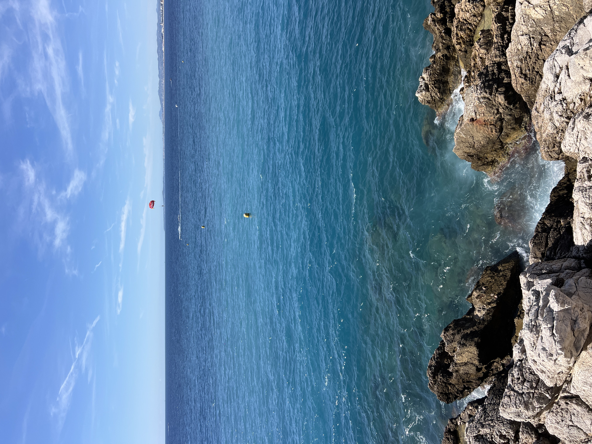 This is a photograph of the Mediterranean Sea and coastline near Nice, France, on a clear, sunny day. The foreground features a rugged, rocky shoreline with large, weathered rocks of varying colors, including white, grey, and brown. Waves are crashing against the rocks, creating white foam.
Beyond the shoreline, the water transitions from a clear, turquoise hue near the coast to a deeper blue further out. Several yellow buoys are visible in the water. In the middle distance, a person is parasailing, attached to a red parachute. There is a boat in the water, likely towing the parasailer, and some distant vessels.
The horizon line is distinct, separating the sea from the sky. The sky is a bright blue with scattered white, wispy clouds. In the far distance, a hazy coastline with mountains and buildings is visible, indicating the presence of land, possibly the city of Nice. The overall impression is of a vibrant, active coastal scene.