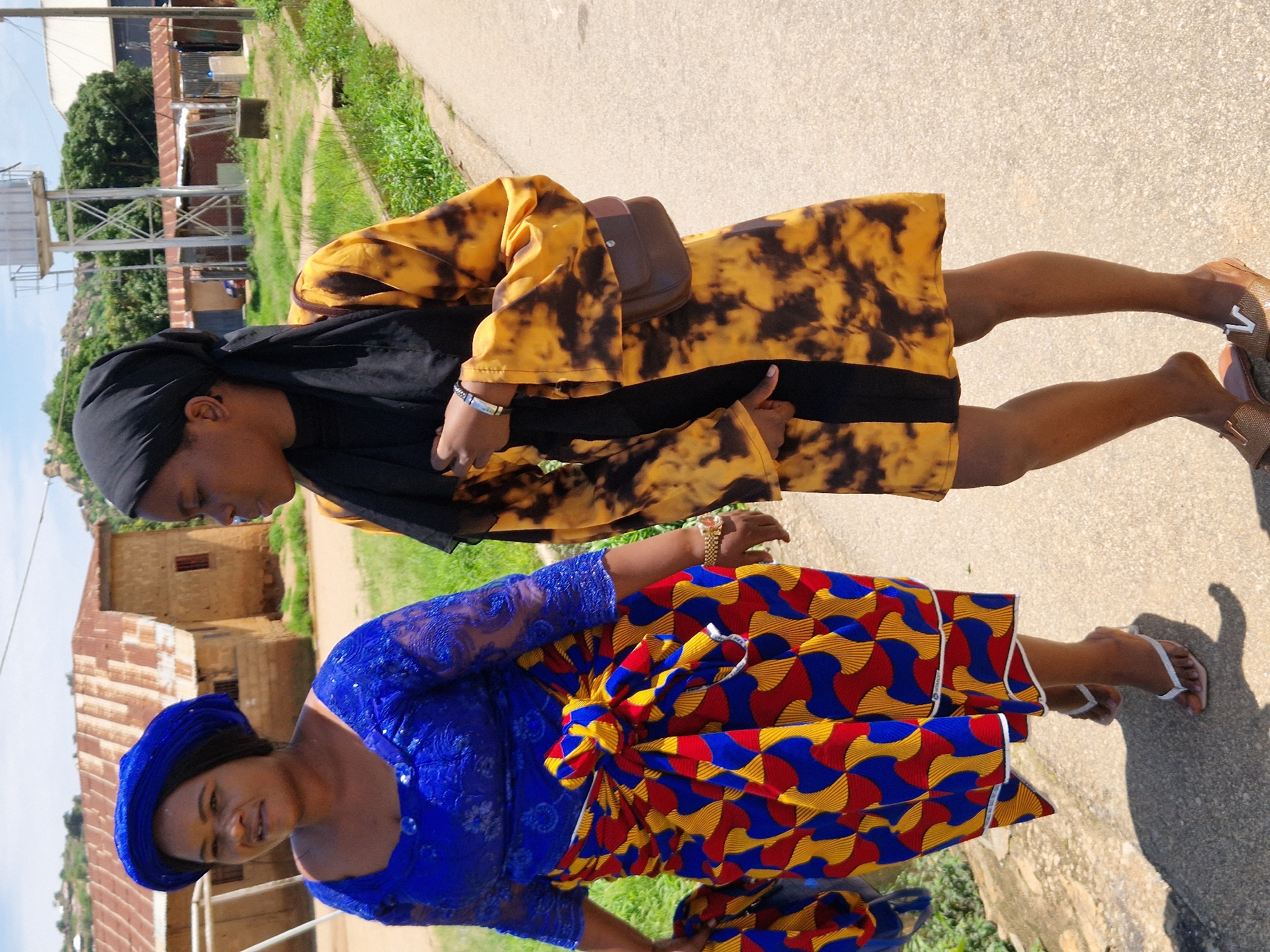 Two women are walking on a paved road in Kabong, Nigeria. The woman on the left is wearing a blue headwrap and a blue lace top with a vibrant Ankara fabric skirt. The woman on the right is wearing a black hijab and a yellow and black tie-dye dress, with a small brown purse slung across her body. Both women are looking forward, suggesting they are walking in the same direction. The background shows a residential area with simple buildings, some with corrugated iron roofs. Vegetation is visible, with grass and trees scattered throughout the scene. The sky is clear and blue, indicating a sunny day.