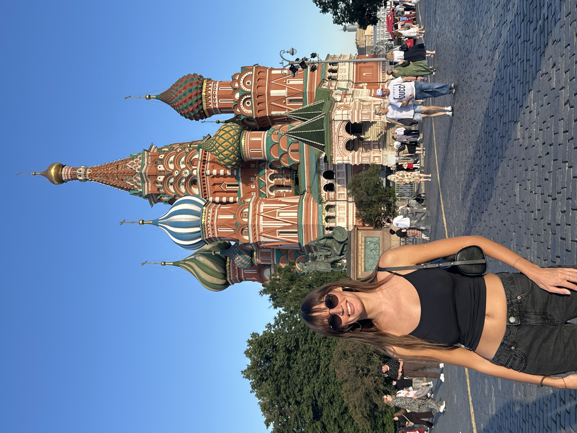 In the foreground, a woman with long brown hair and sunglasses stands facing the camera, wearing a black tank top and dark jeans. She is smiling and carrying a black purse. Behind her, Saint Basil's Cathedral, a Russian Orthodox church known for its colorful onion domes, dominates the scene. The cathedral is a vibrant red with green and blue accents. People are walking in the background on a cobblestone plaza. The sky is clear and blue.