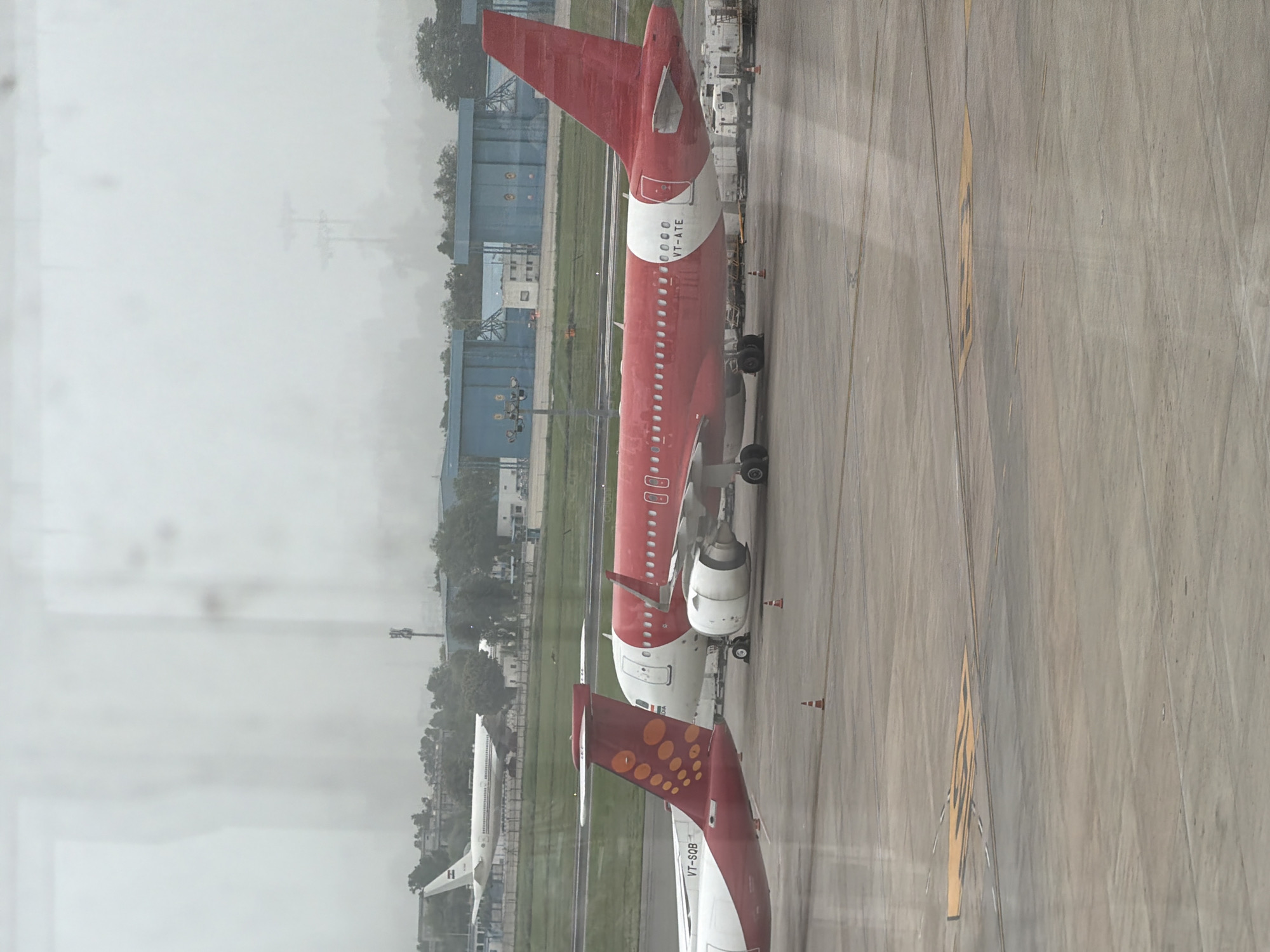 The image shows an airport tarmac on a cloudy day, likely taken from inside an airport terminal looking out a window. In the foreground, two airplanes are parked. The plane on the right is primarily red with a white stripe around the rear fuselage, featuring the registration VT-ATE. The plane on the left is white with a red tail featuring an orange circular pattern and the registration VT-SQB. In the background, a white airplane is visible on the runway, and beyond that, there are airport buildings and trees. The tarmac is marked with yellow lines and cones. The overall atmosphere is overcast, suggesting potential rain or fog. The location is identified as Indira Gandhi International Airport (DEL) in New Delhi, India, based on the context of airplanes and airport infrastructure.