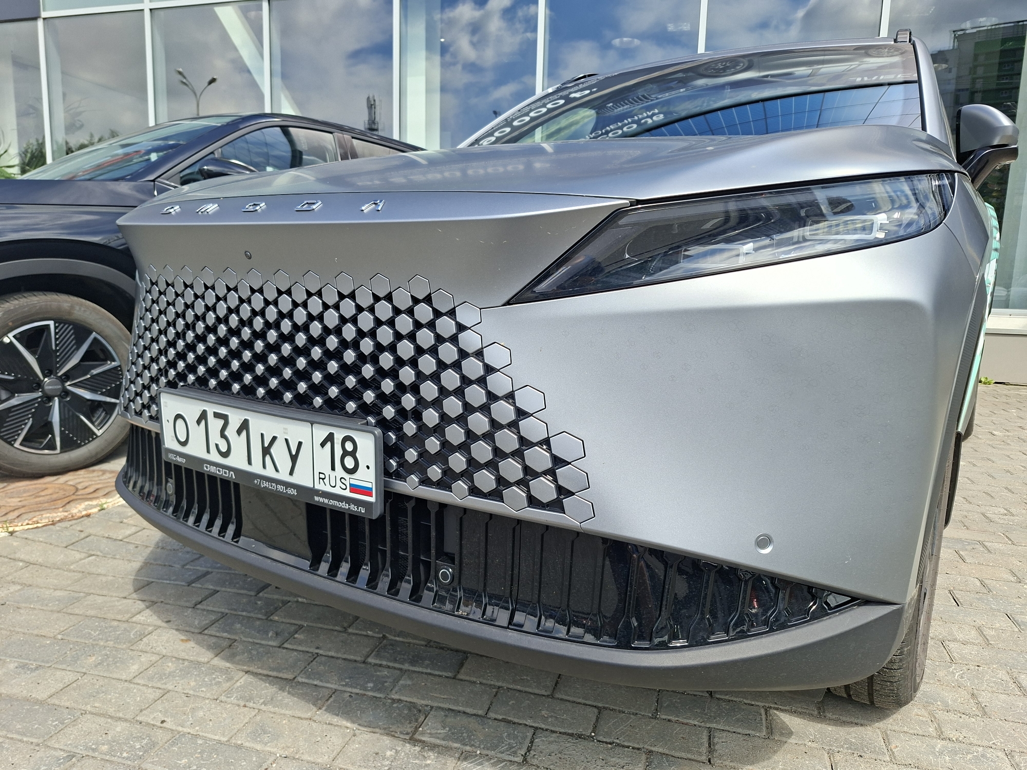 The image shows the front of a silver OMODA car parked on a paved surface, likely in front of a modern building with large windows reflecting the sky. A dark-colored car is partially visible to the left. The license plate of the OMODA car reads "0 131 KY 18", indicating it is registered in the Udmurt Republic, Russia, which includes the city of Izhevsk. The OMODA logo is visible on the car's grille, which features a distinctive honeycomb pattern. A parking sensor is visible on the bumper. The overall environment suggests a car dealership or a public parking area in a city.