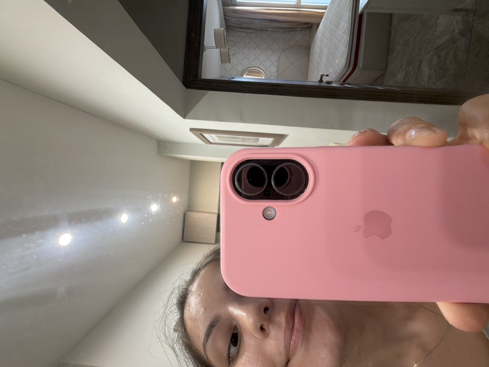 The image is a selfie taken in a hallway. The subject, a young woman with dark hair, is holding a pink smartphone in front of her face, obscuring a portion of her facial features. The phone has a dual-camera setup and an Apple logo on its back. In the background, a doorway leads to a bedroom with a bed, a nightstand, and a window with curtains. The hallway itself is lit by ceiling lights and has a framed picture on the wall. The location is identified as Burgas, Bulgaria.