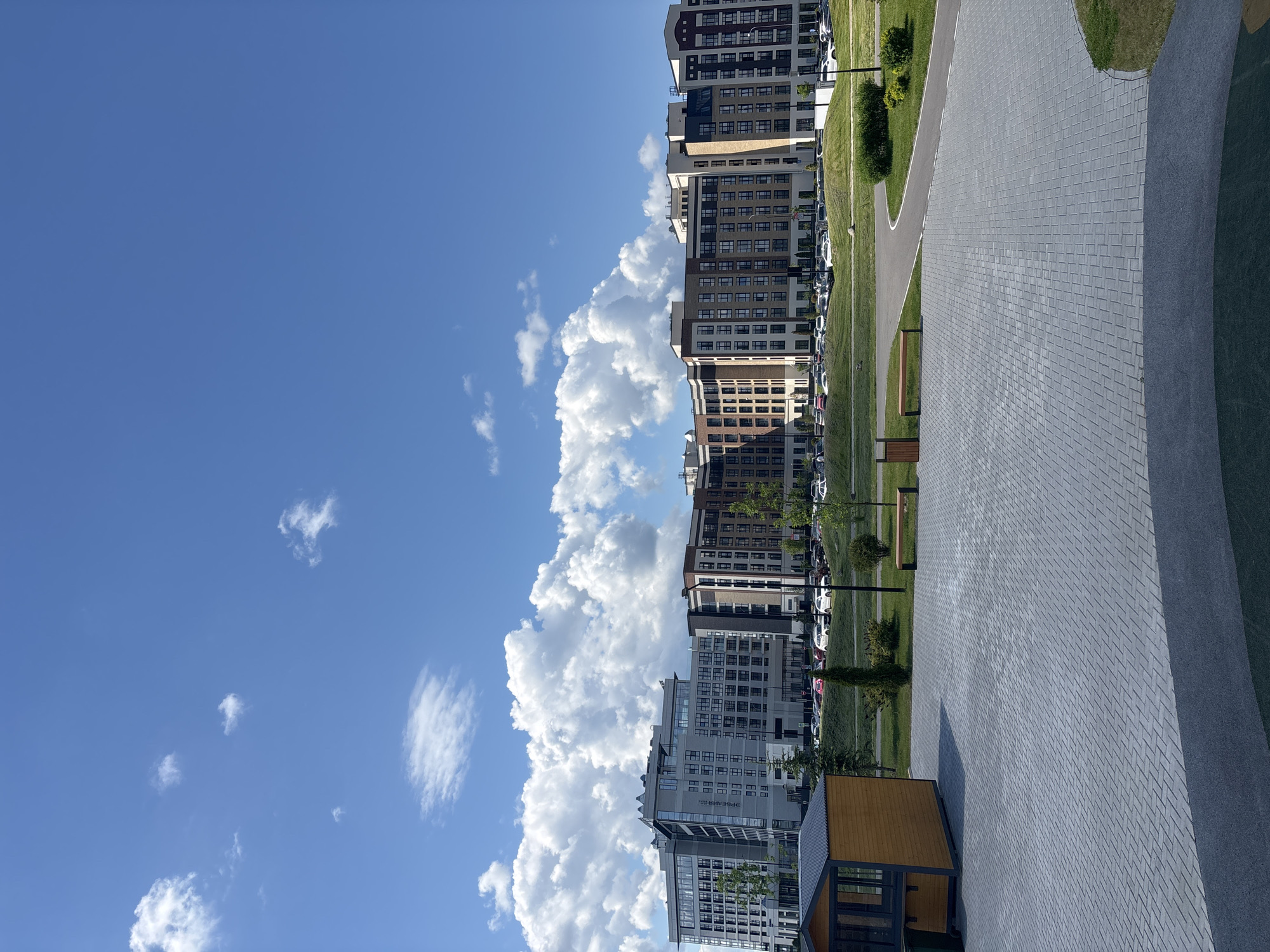 The image shows a view of a street in Varaksino, Russia. The sky is blue with white cumulus clouds. Several modern apartment buildings line the street. There is a paved walkway on the right side of the image, and grass and trees are visible between the walkway and the buildings. Cars are parked along the street. A small, wooden structure is visible in the bottom right corner of the image. The image is taken from a ground-level perspective.
